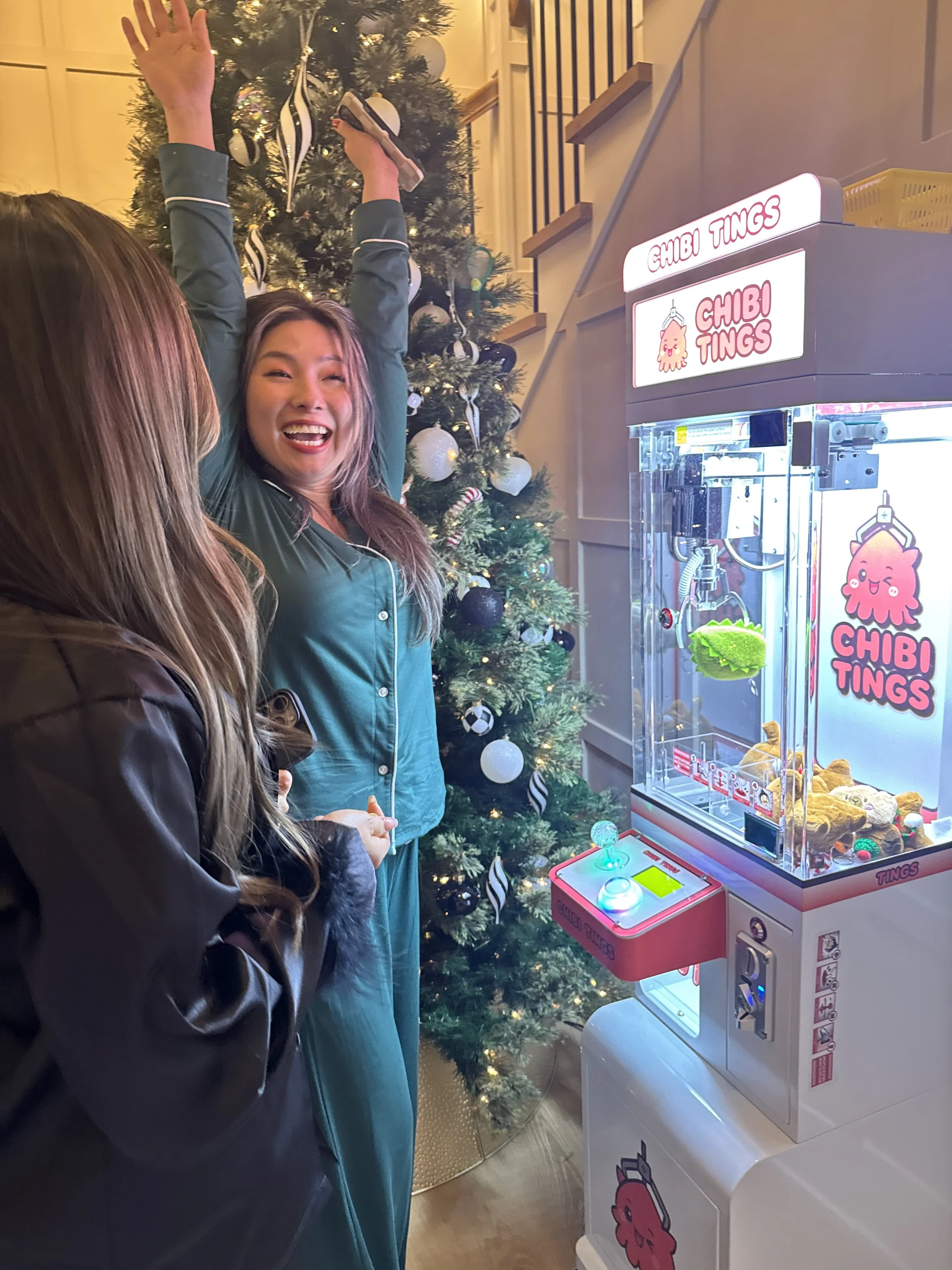 Two women smiling, one in pajamas and another in a jacket, appear to be celebrating near a Christmas tree and a claw machine decorated with pink and white accents labeled 'Chibi Tings'.