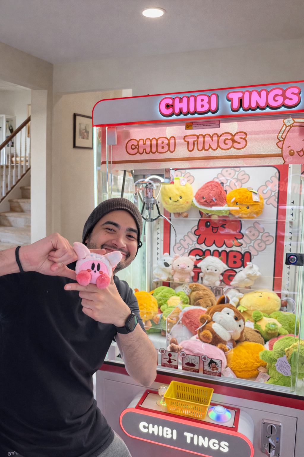 A smiling man wearing a black shirt and a gray beanie posing in front of a claw machine filled with kawaii plush toys, including characters from Cinnamoroll and Kirby.