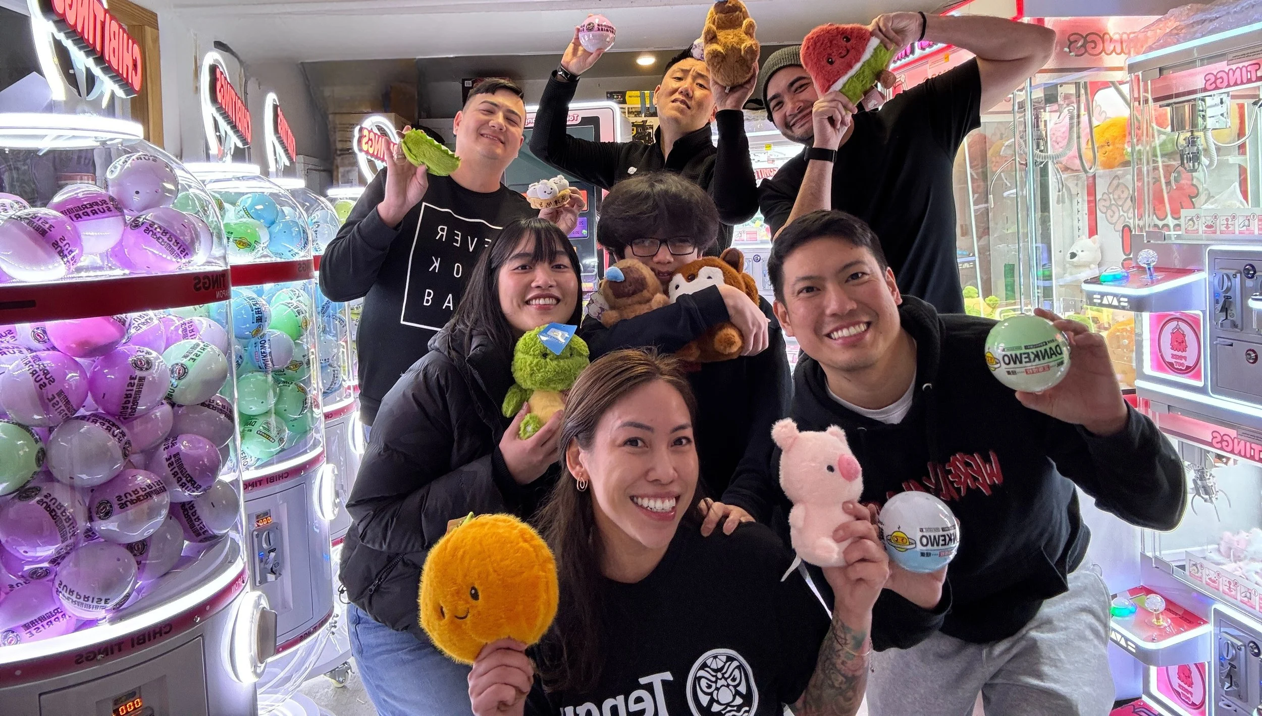 Group of smiling young adults at an arcade holding plush toys and prizes, surrounded by claw machine prize displays.