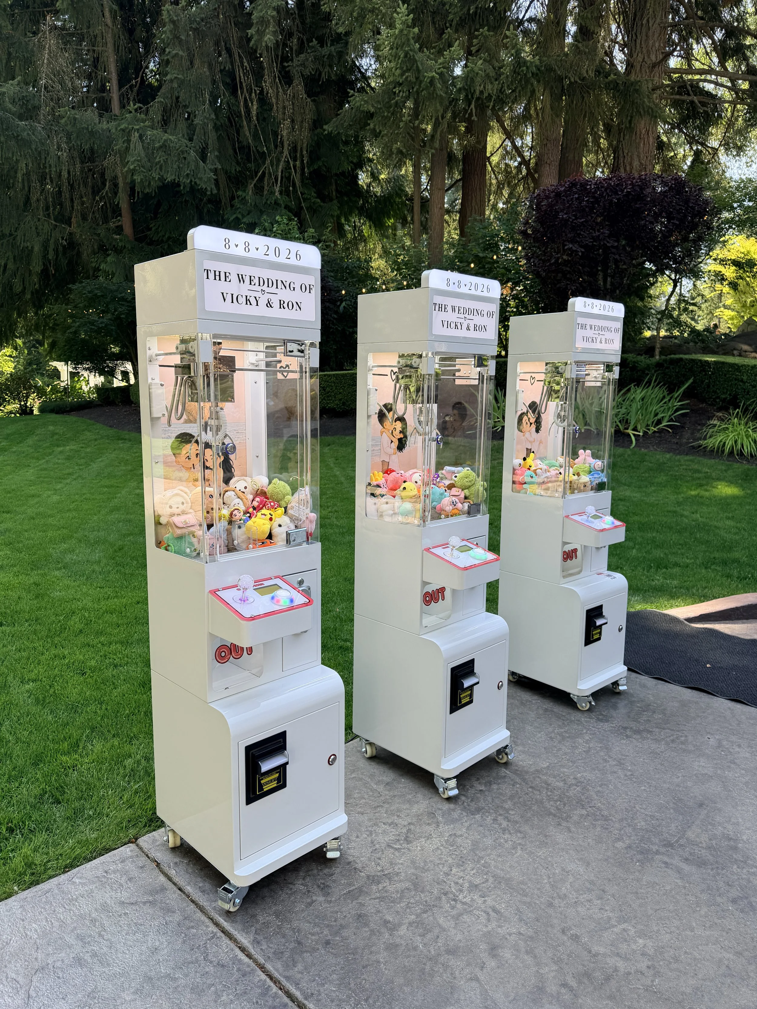 Three white claw machines filled with plush toys, placed outdoors on a sidewalk on a grassy area with trees in the background, and a black mat on the ground near the machines.