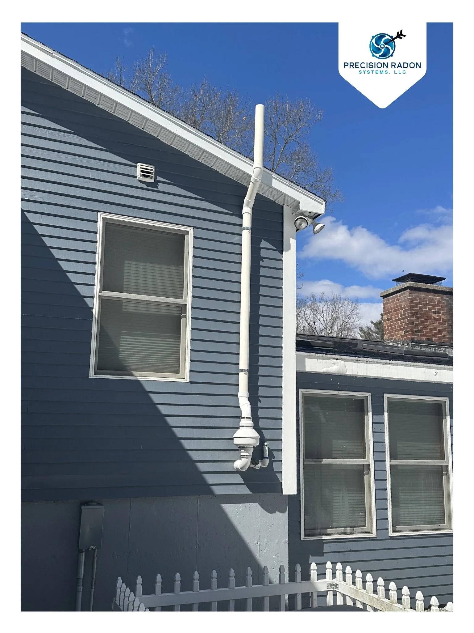 Side of a blue house with three double-pane windows, a white gutter system, and a vertical pipe connected to a rain downspout. There is a white fence in the foreground and a brick chimney in the background under a blue sky with some clouds.