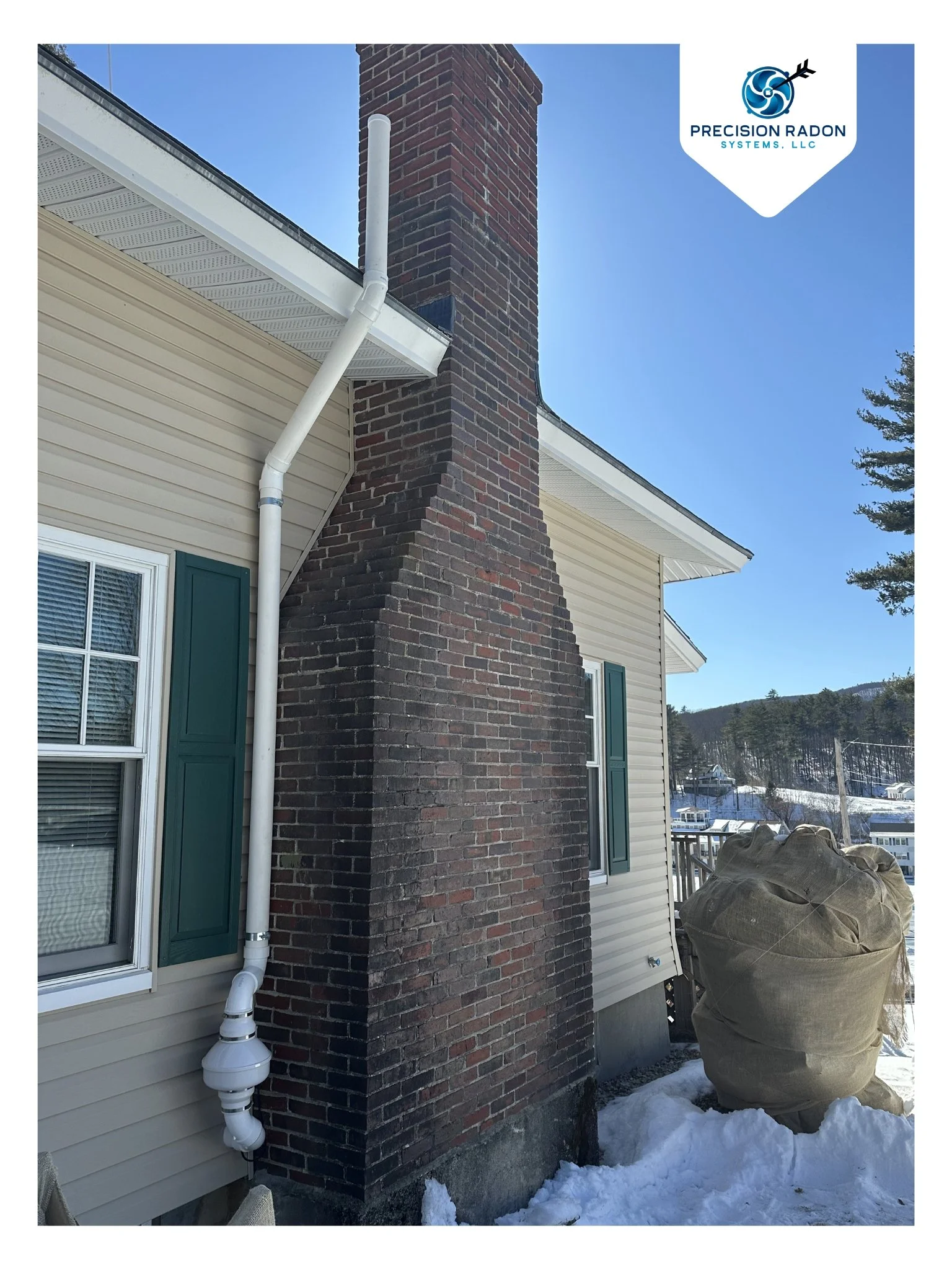 Close-up of a house exterior with a brick chimney, white vent pipes, green shutters, and snow on the ground, with a clear blue sky and distant hills.