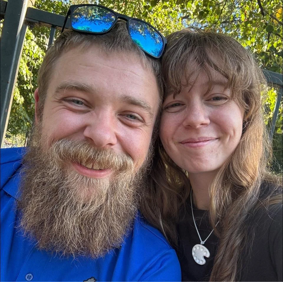 Close-up of a smiling man with a beard and blue shirt, and a woman with wavy brown hair and a shell necklace, outdoors with trees and sunlight in the background.