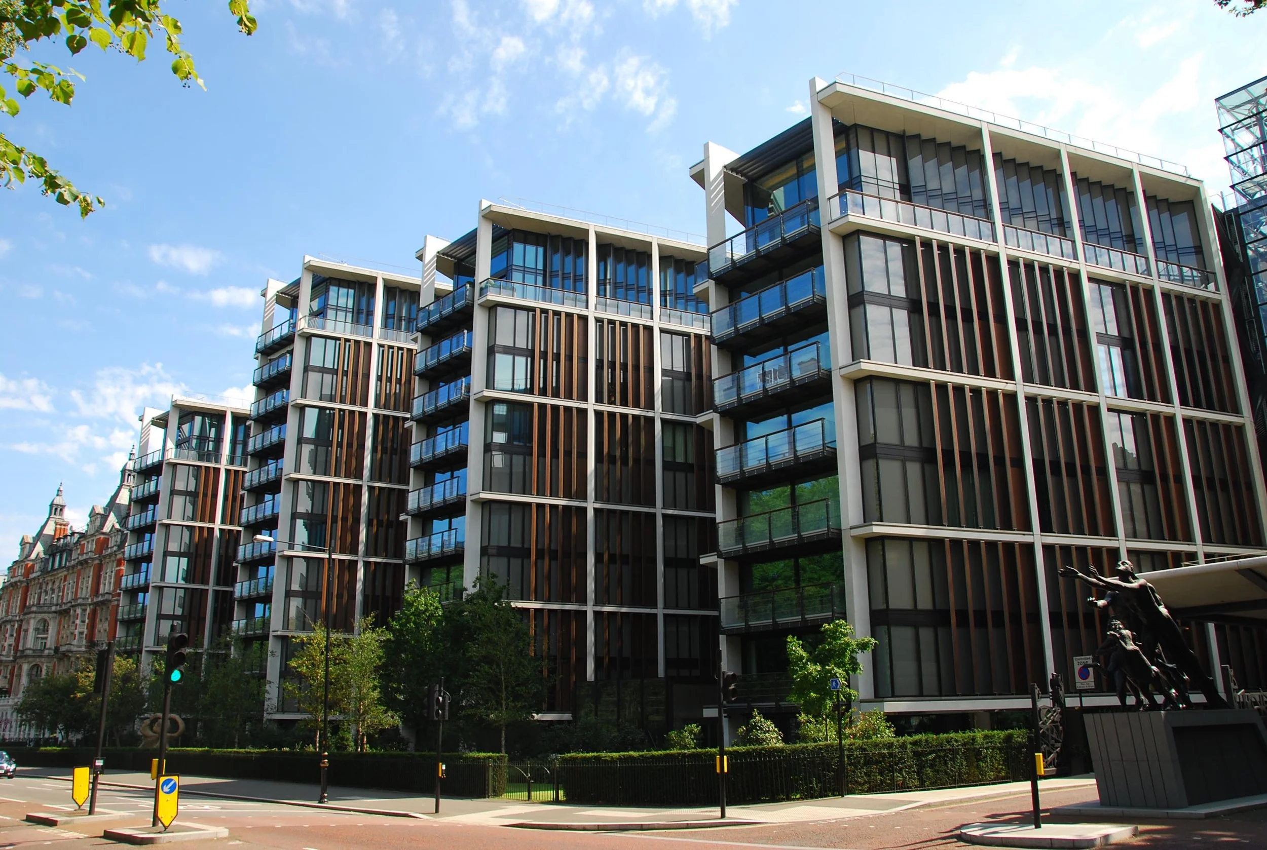 Modern multi-story apartment building with glass windows and balconies, situated on a city street with trees and streetlights in the foreground.