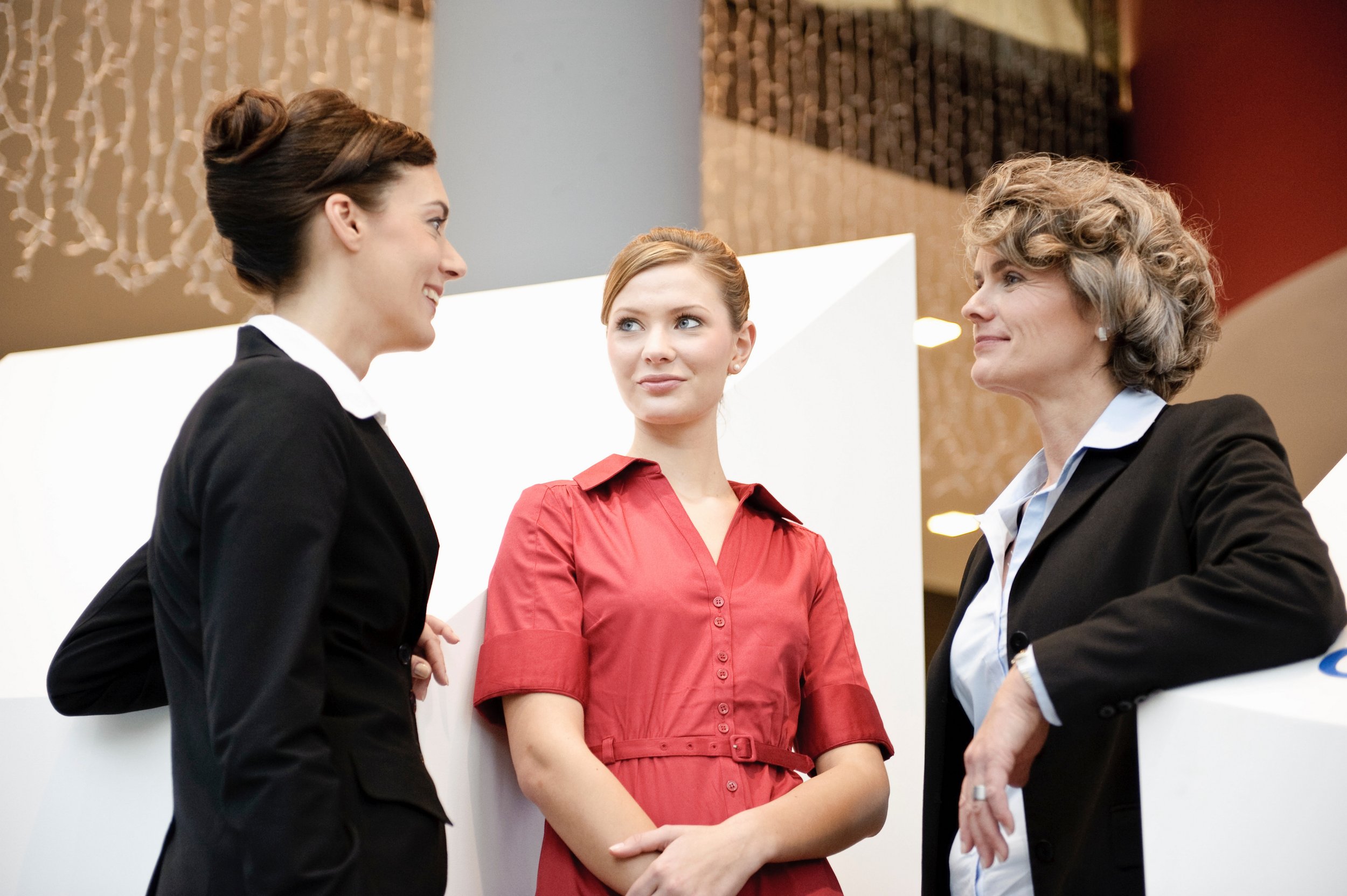 Three women engaged in conversation, one in a black suit, another in a red dress, and the third in a black suit, inside a modern office or conference space.