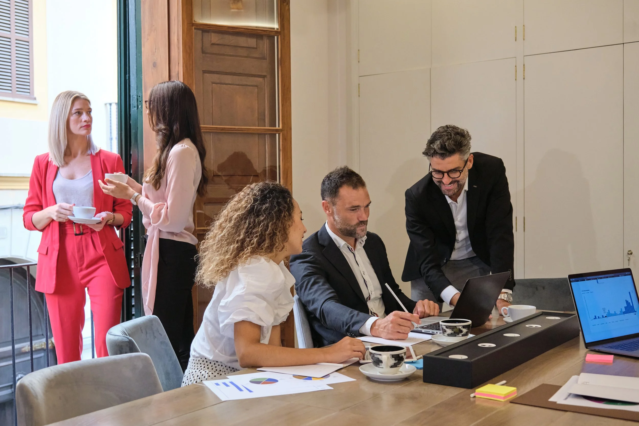Five professionals in a meeting room, three sitting at a table with laptops and documents, two women standing near a door with coffee cups, engaging in conversation.