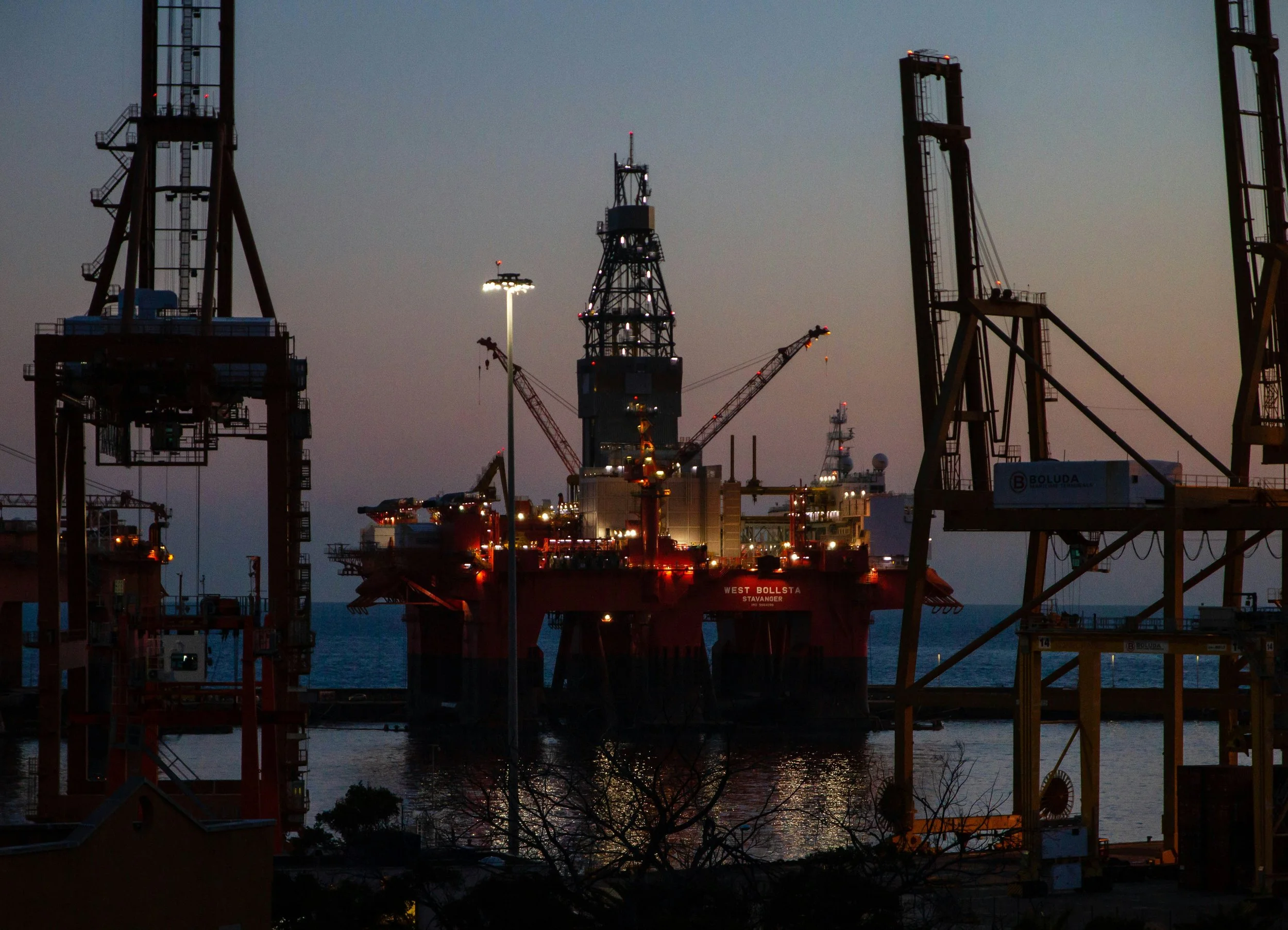 Offshore oil drilling rig at dusk surrounded by construction cranes and dockyard structures.