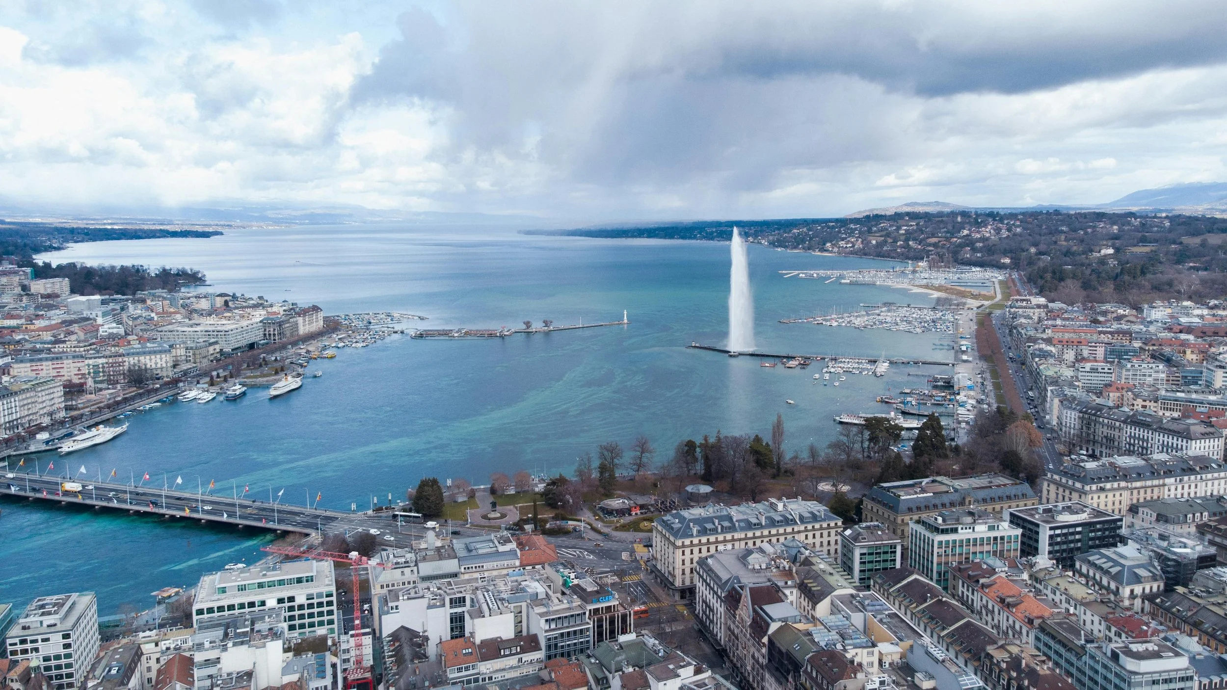 Aerial view of a city with a large body of water, a prominent fountain shooting water into the air, boats in the harbor, and a bridge crossing the water. Urban buildings and streets are visible in the foreground.