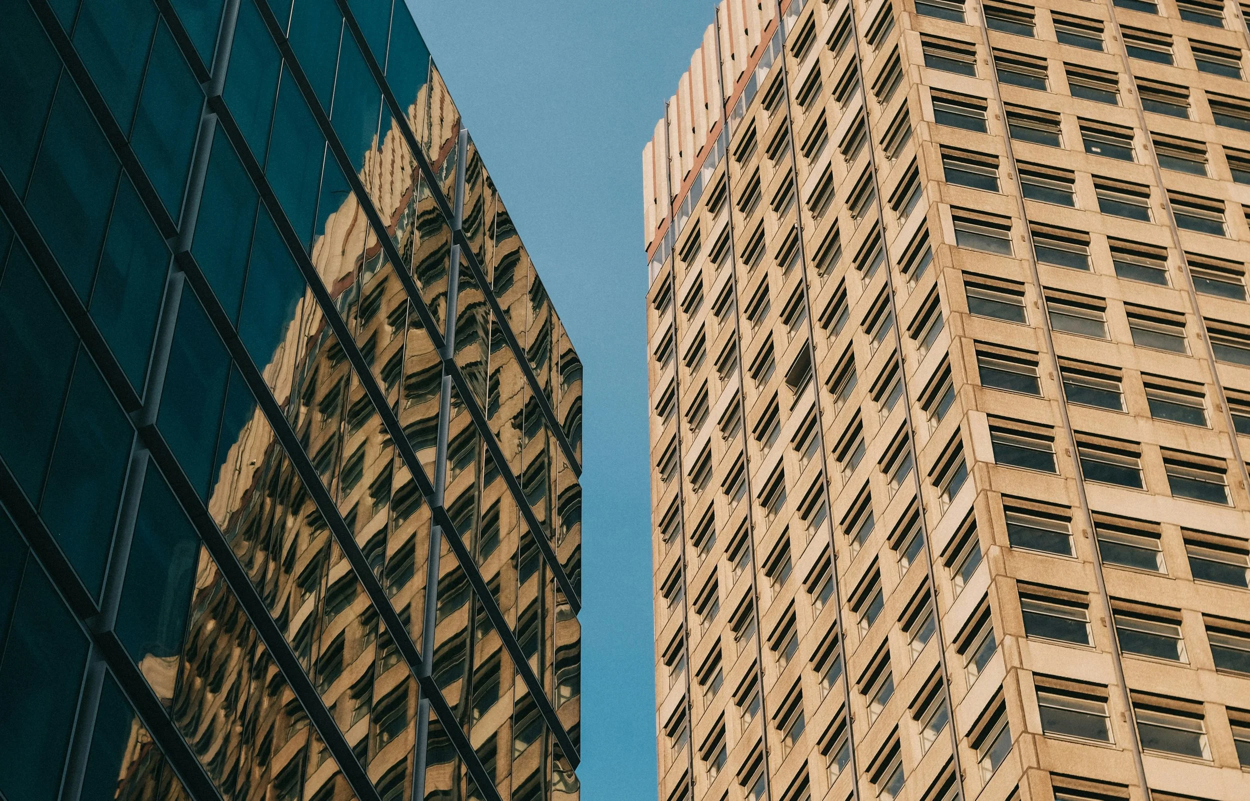 Looking up at two tall buildings with clear blue sky in the background, one building with reflective glass windows and the other with beige stone facade and numerous rectangular windows.