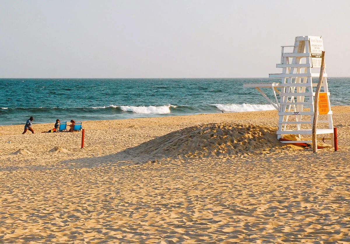 Empty sandy beach with a white lifeguard tower on the right and three people sitting on chairs with a child nearby on the left, with waves breaking in the ocean in the background.