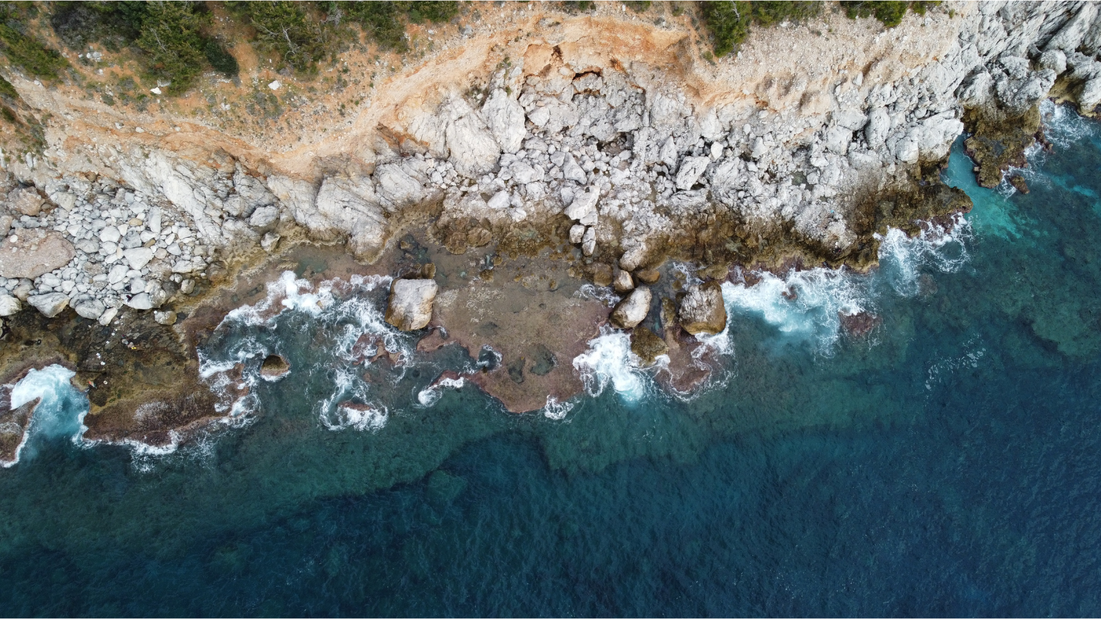 Aerial view of a rugged coastline with rocky cliffs and clear blue ocean water crashing against the shore.