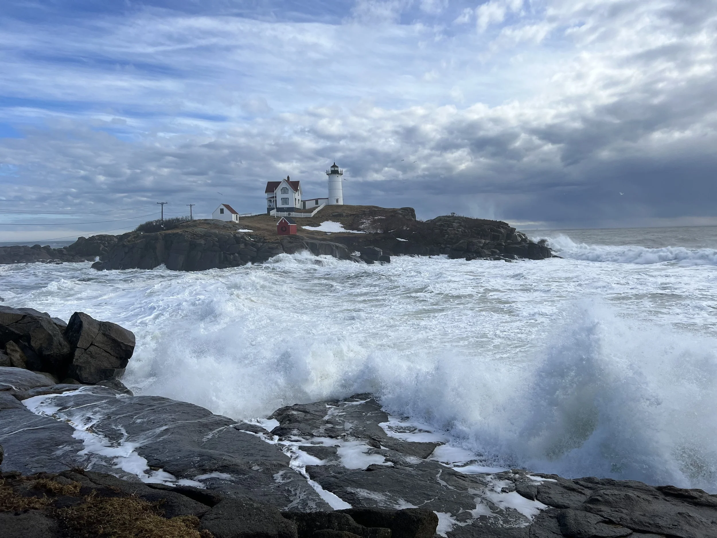 Ocean waves crashing against rocks with a lighthouse and house on a cliff in the background under partly cloudy sky.