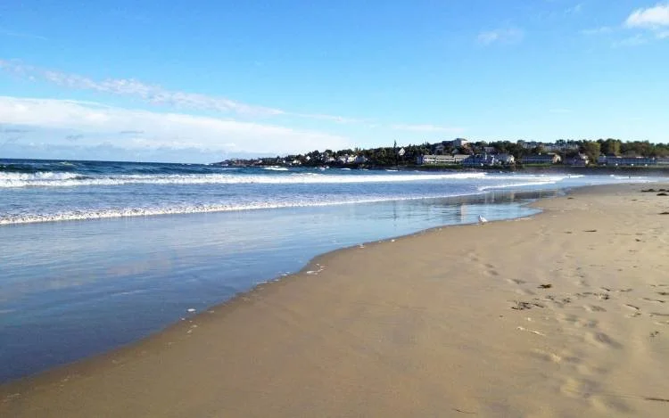 Sandy beach with gentle waves and a distant community along the shoreline under a partly cloudy blue sky.