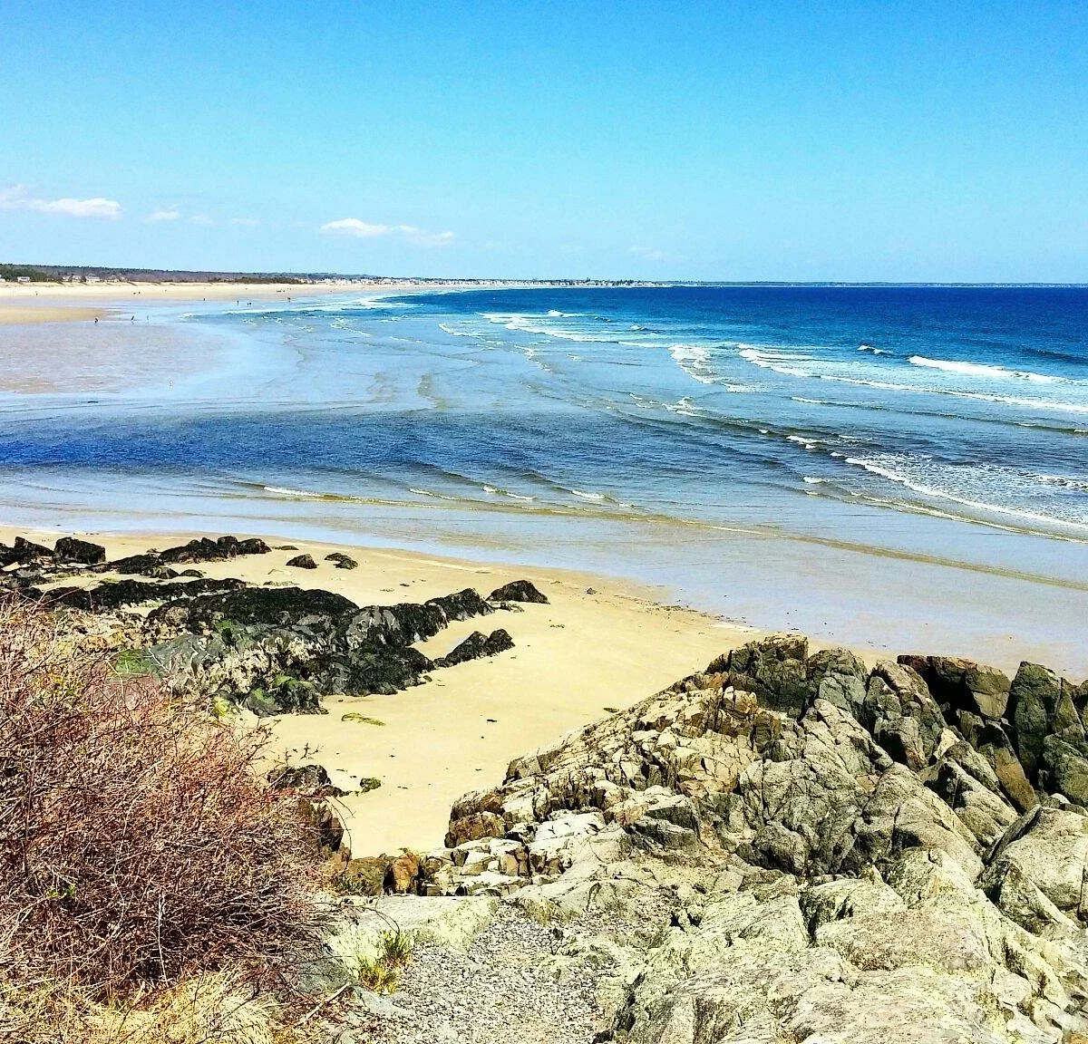 View of a sandy beach with rocks, gentle waves, and a clear blue sky.