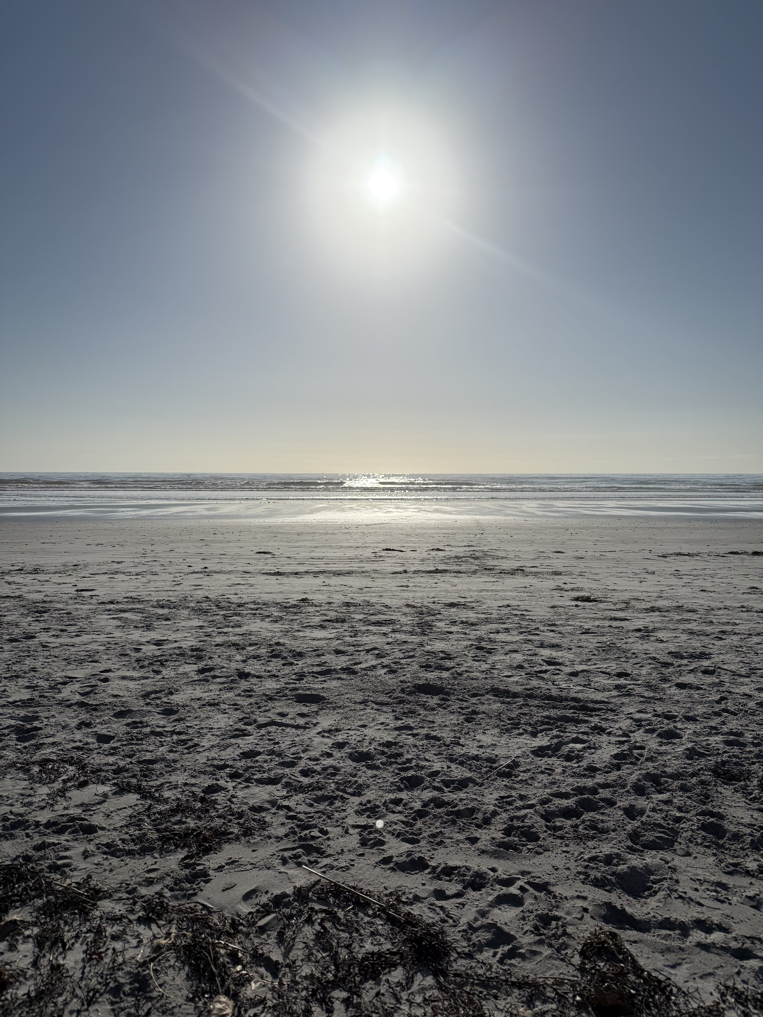 A sunny beach with a clear sky, sandy shore, gentle waves, and sunlight reflecting on the water.