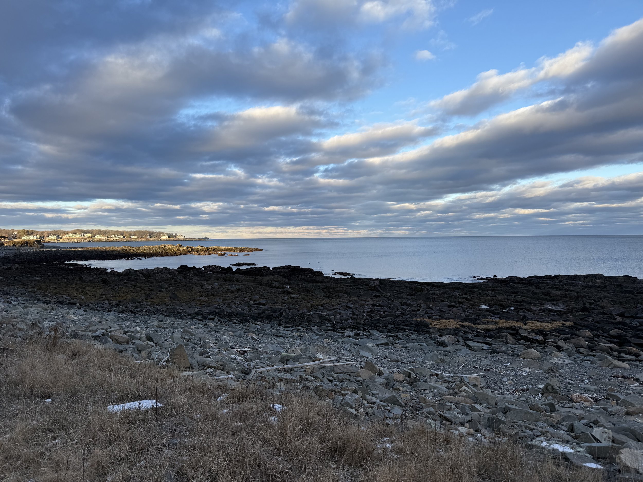 View of a rocky shoreline by the ocean with clouds in the sky and a distant landmass or town.