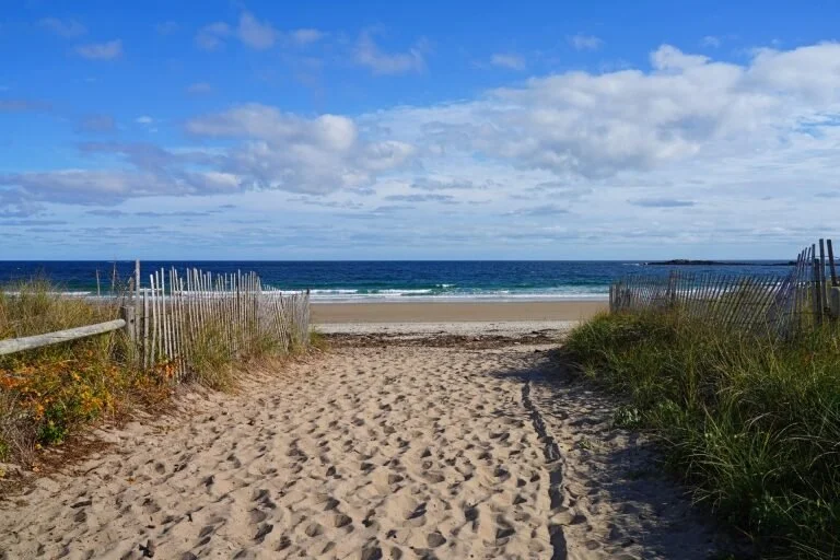 Sandy beach entrance with wooden fences on both sides leading to the ocean, under a partly cloudy sky.