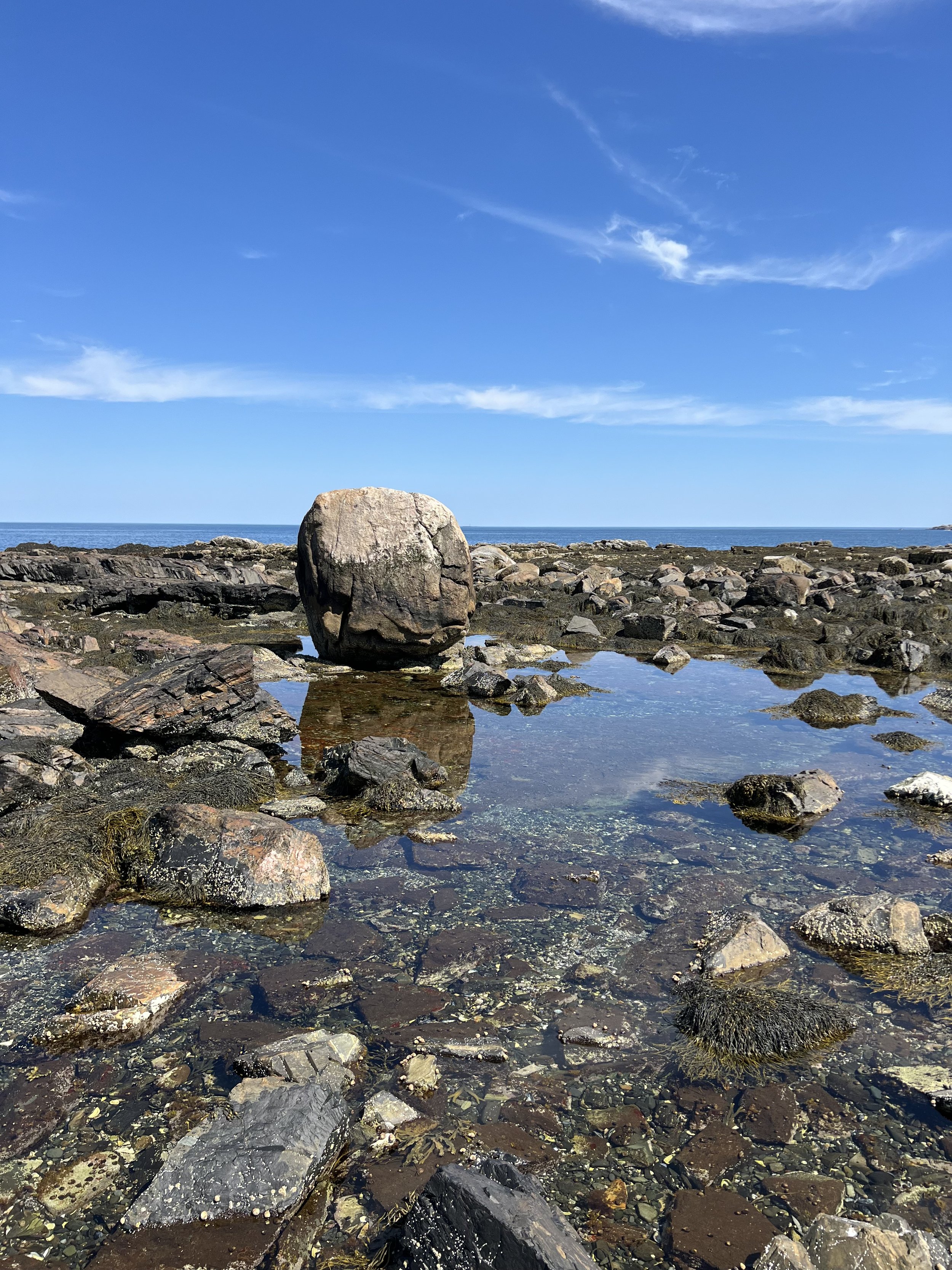 A rocky shoreline with large boulders, some rocks partially submerged in tide pools, under a blue sky with wispy clouds.