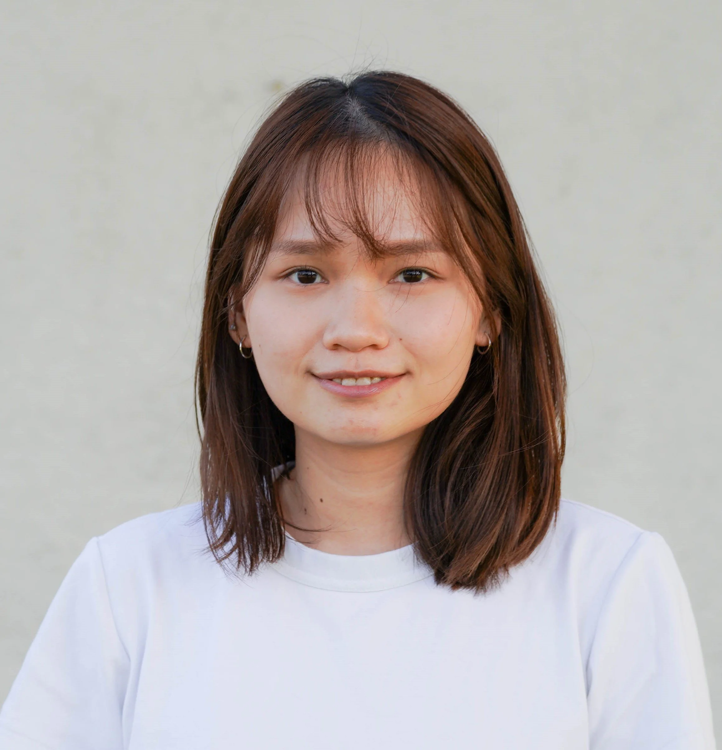 A young woman with brown hair and small hoop earrings smiling in front of a plain light-colored background.