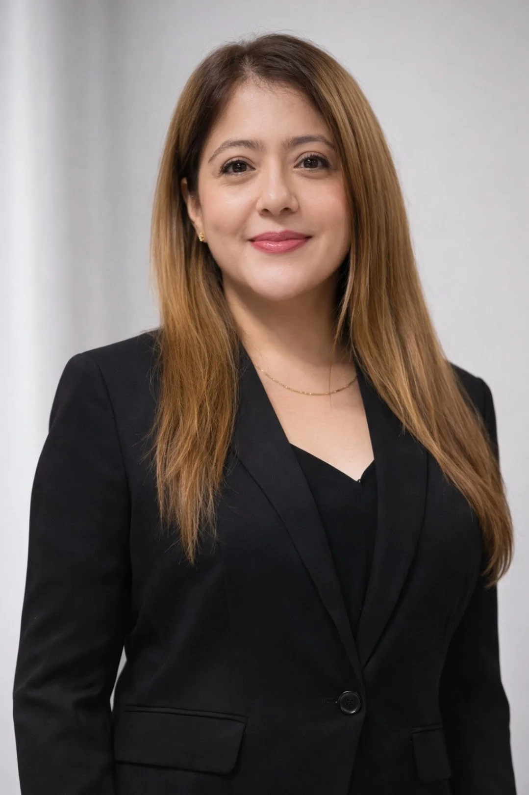 A woman with long, light brown hair wearing a black blazer and black top, smiling at the camera, standing in front of a plain, light gray background.