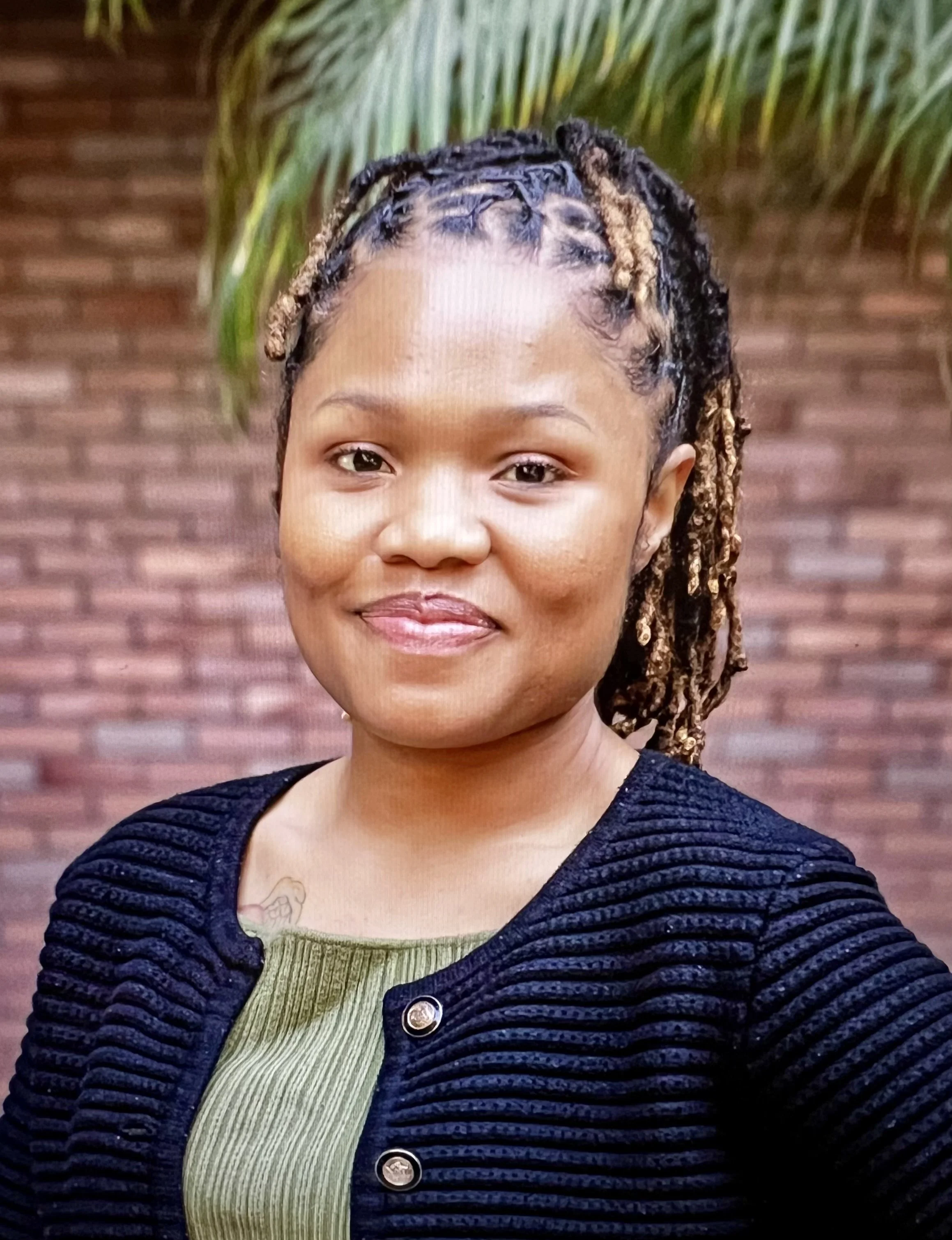 A woman smiling outdoors in front of a brick wall and green foliage, wearing a black ribbed jacket and an olive green top.