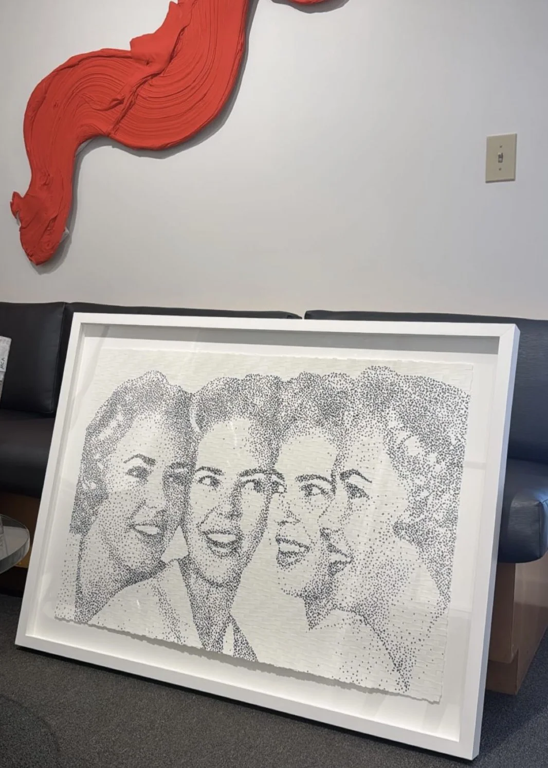 A framed black-and-white photo mosaic of four women with curly hair, smiling and looking to the left, placed on the floor leaning against a sofa in a room with a white wall.