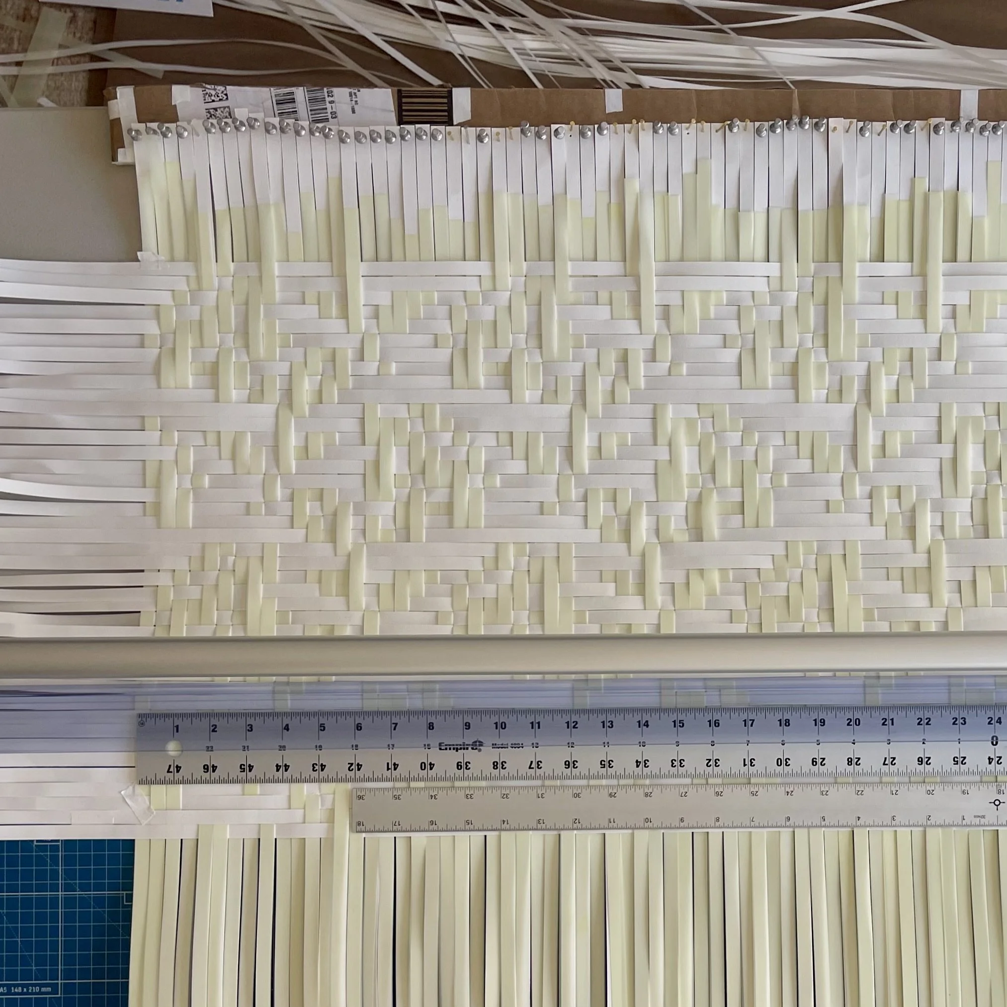 A collection of white and yellow paper strips arranged in a woven pattern, with a ruler at the bottom and a blue grid cutting mat at the lower left corner.