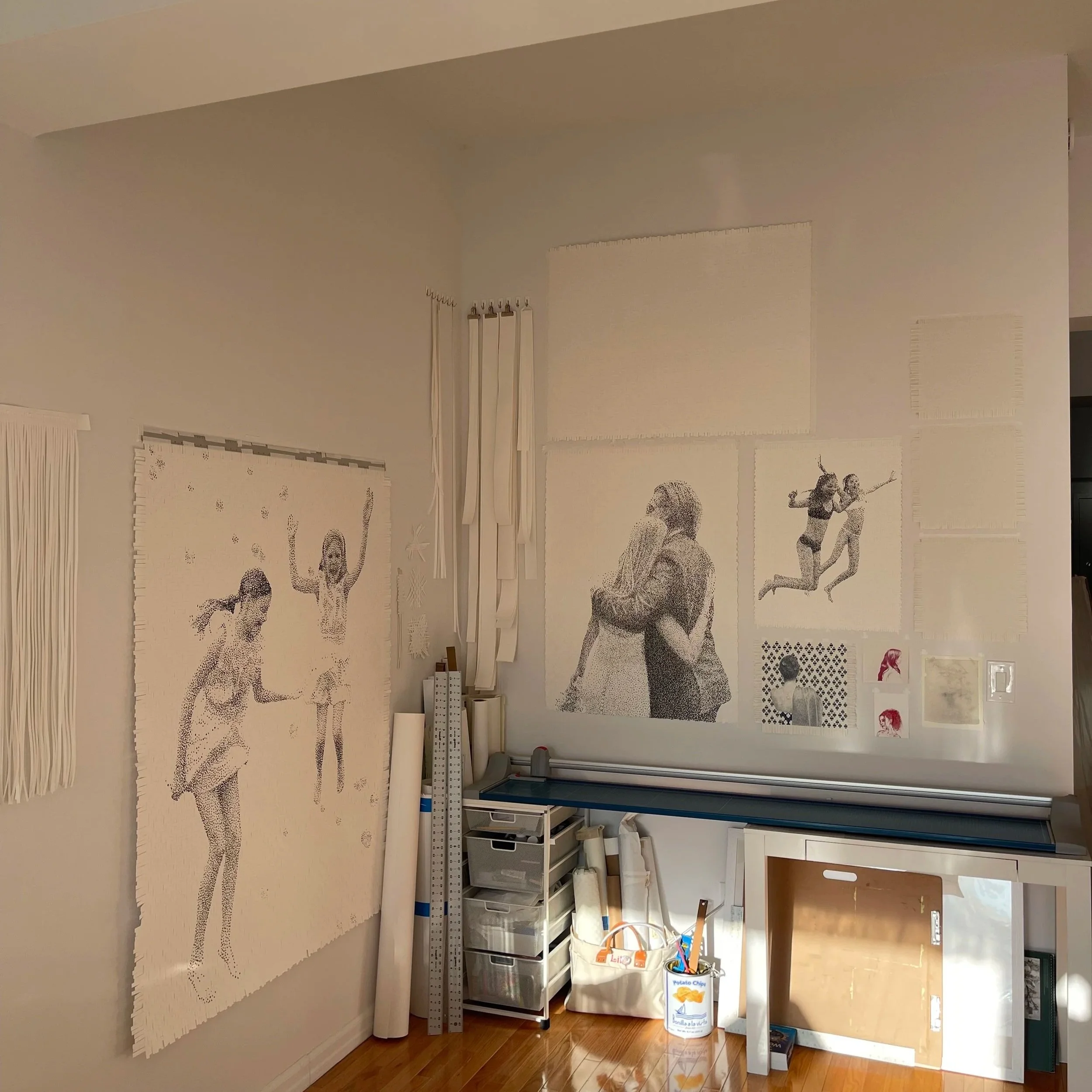 Wall with various mosaic art pieces, including images of people, including two children, two women, and a man hugging a woman, with some art pieces still in progress. Art supplies and equipment are on a table and shelves below.