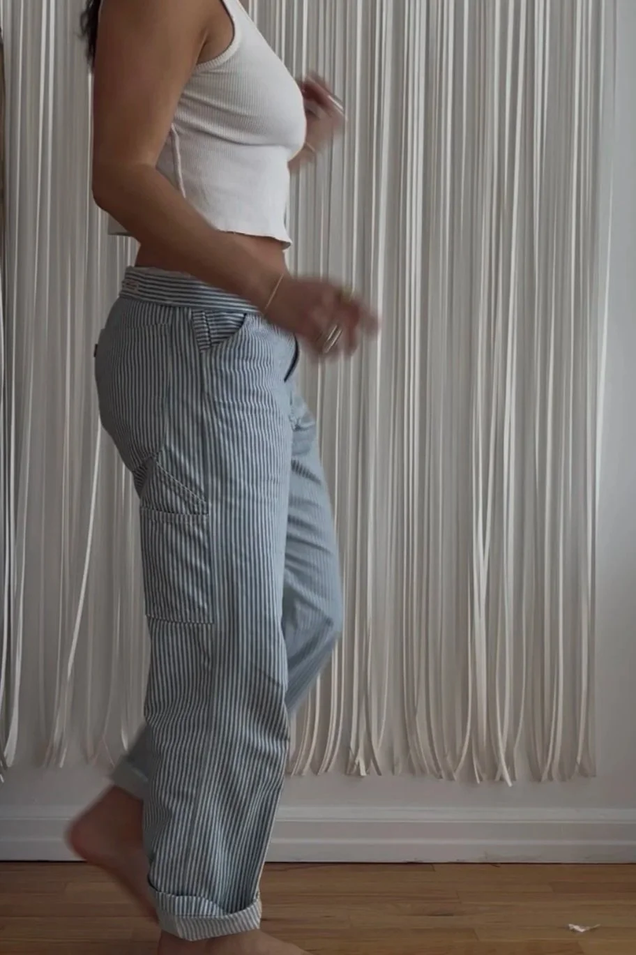 A woman standing in front of cream-colored curtains, wearing a white sleeveless top and striped pajama pants, with one foot slightly raised and holding a small object.