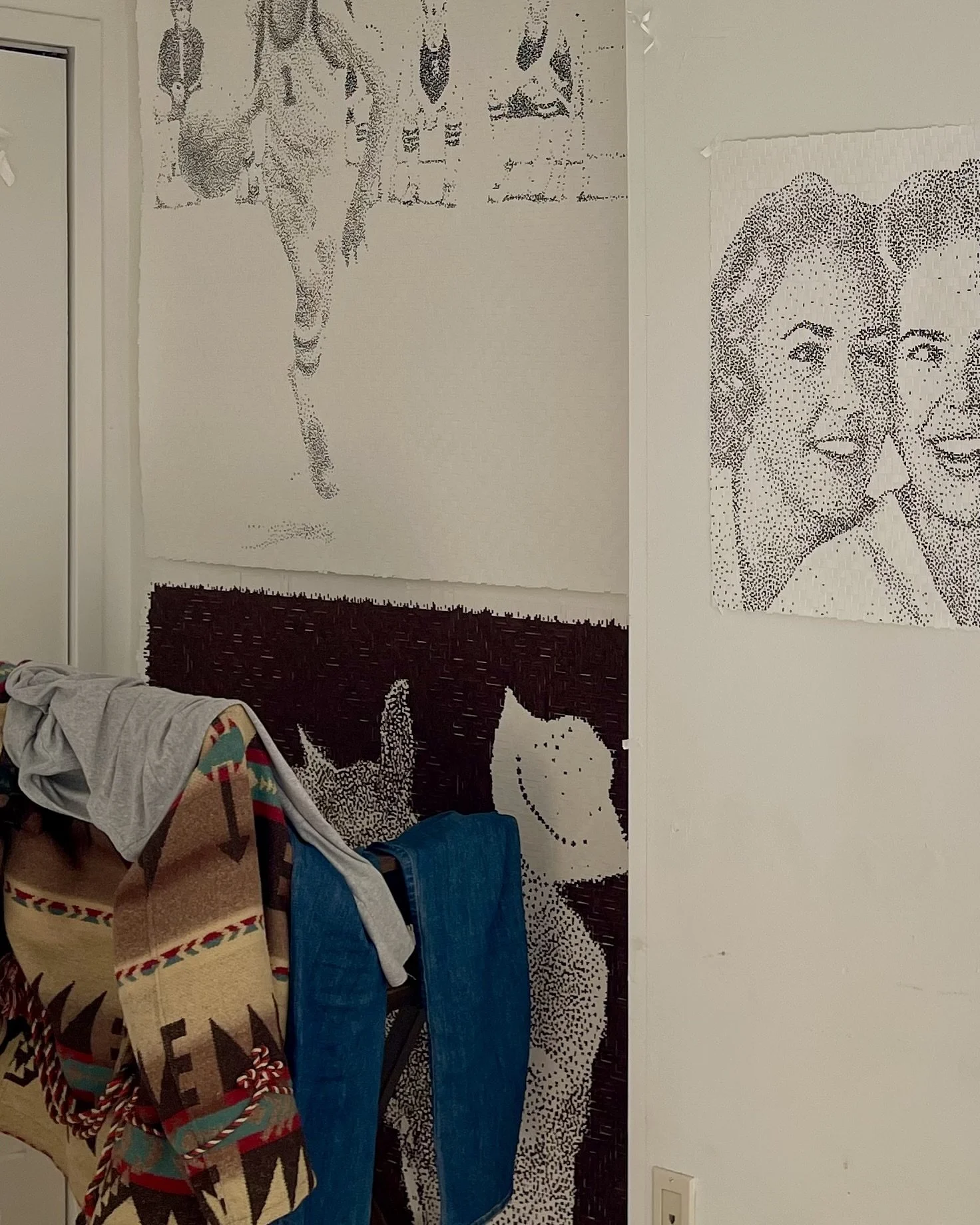 Part of a room with pixelated black and white posters on the wall, including a close-up of two smiling women and a running horse, with laundry hanging in front.