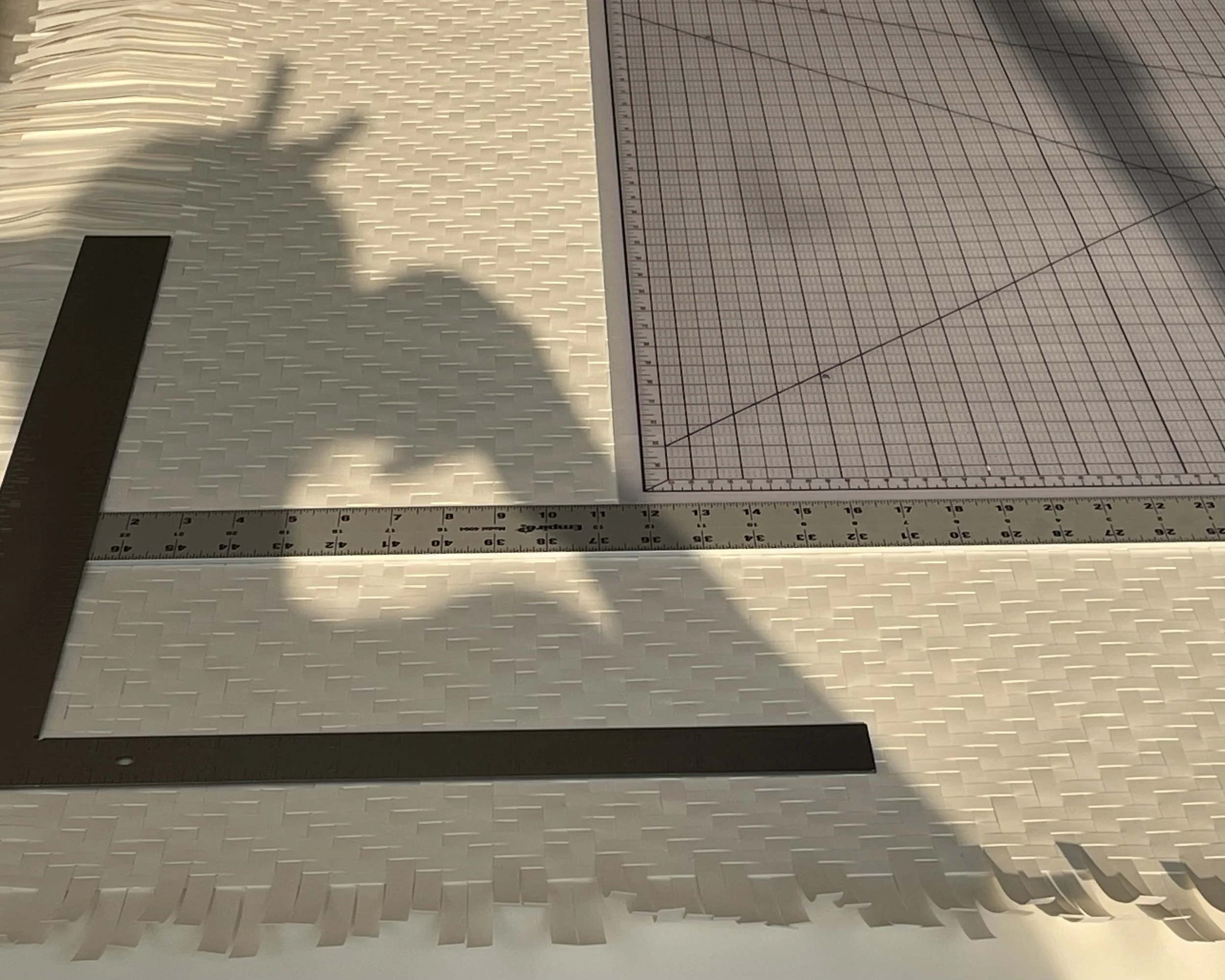 A white textured surface with a metal L-shaped ruler, a black measuring tool, and a sheet of graph paper with grid lines and a diagonal line. Shadows of a plant or branch are cast on the surface.