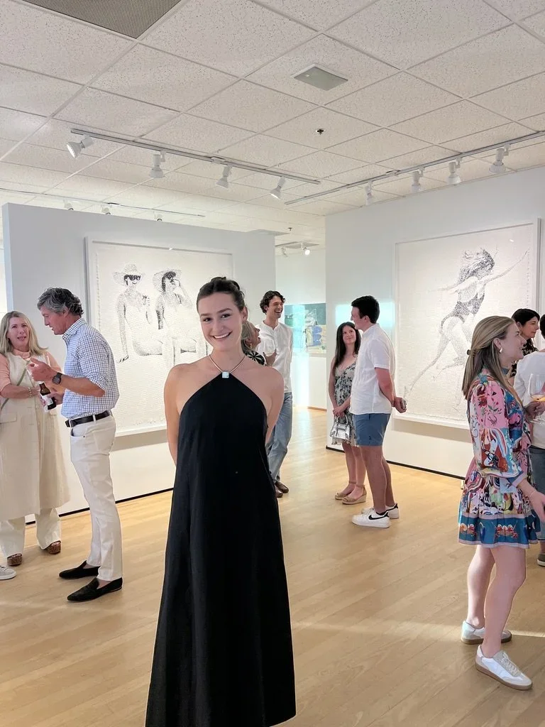 A woman in a black dress standing in an art gallery with other people looking at artwork and talking.