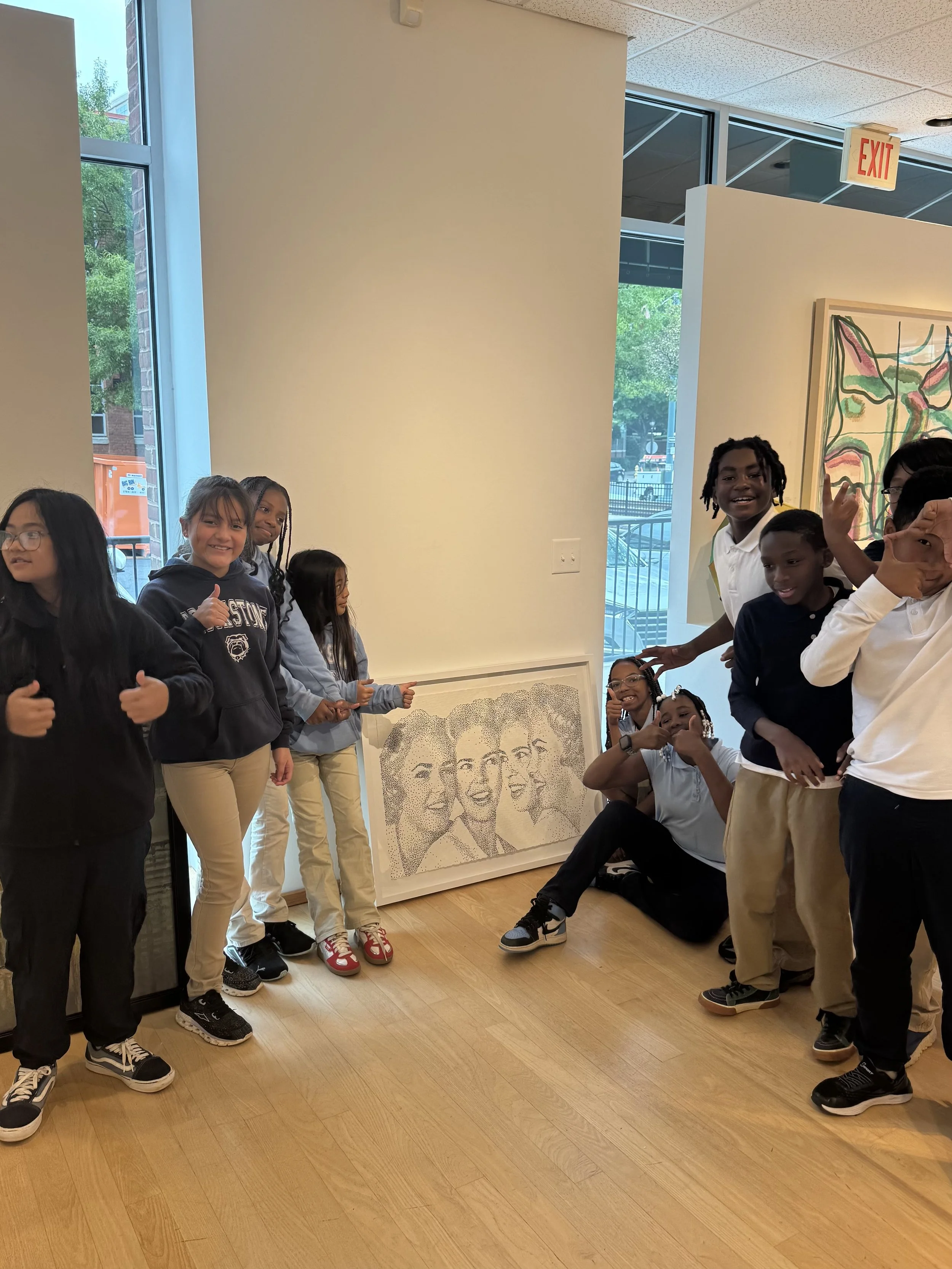 A group of children in an art gallery standing and sitting around a framed artwork that depicts four women's faces. Some of the children are smiling and making gestures.