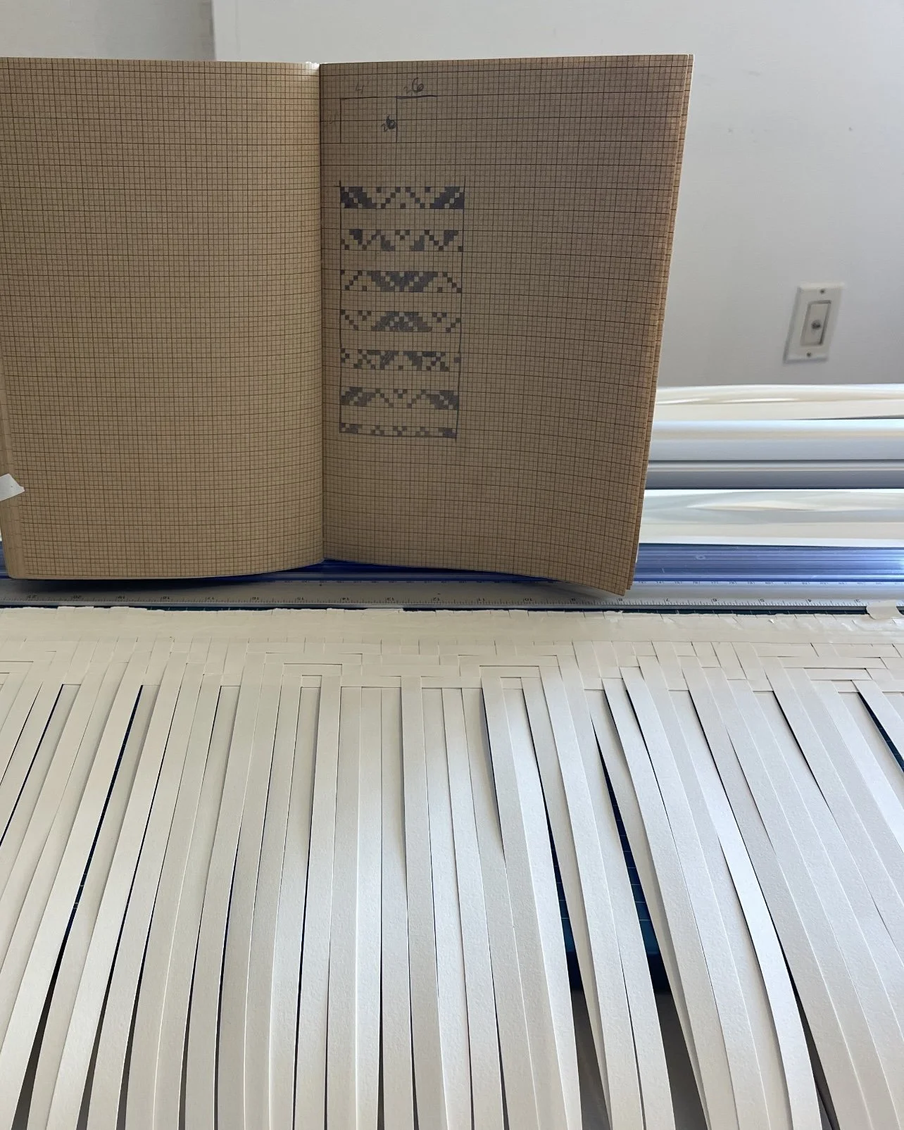 Open grid paper notebook with gray pixel art pattern and pencil sketches, resting on a cutting mat with white strips, near a wall with an electrical outlet.