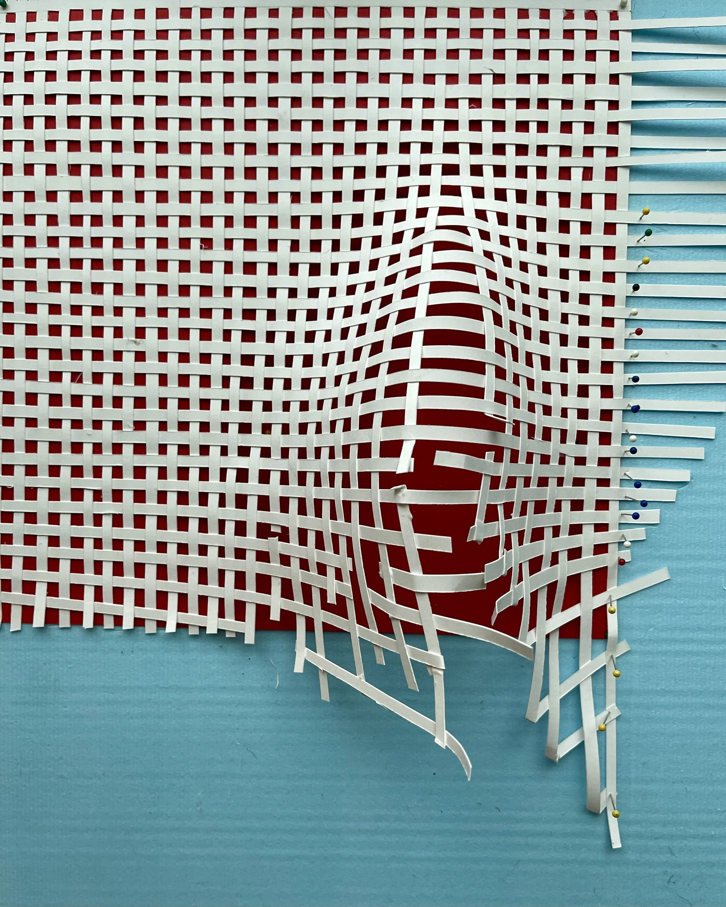 Unfinished paper weaving project with white strips on a red and blue background, held together with pins.