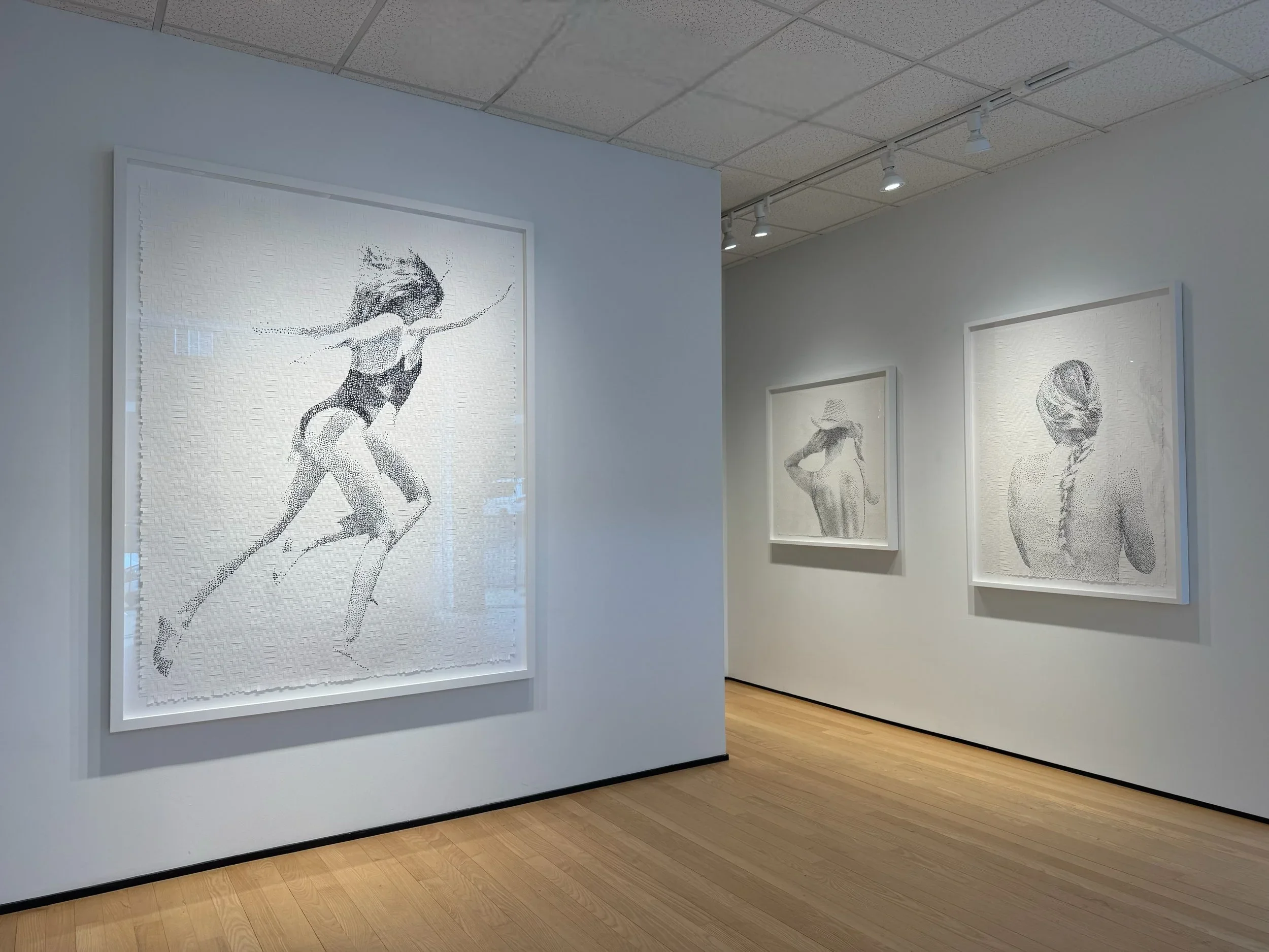Art gallery with three large black-and-white drawings of women on white walls and light wooden floors, ceiling lights illuminating the artwork.