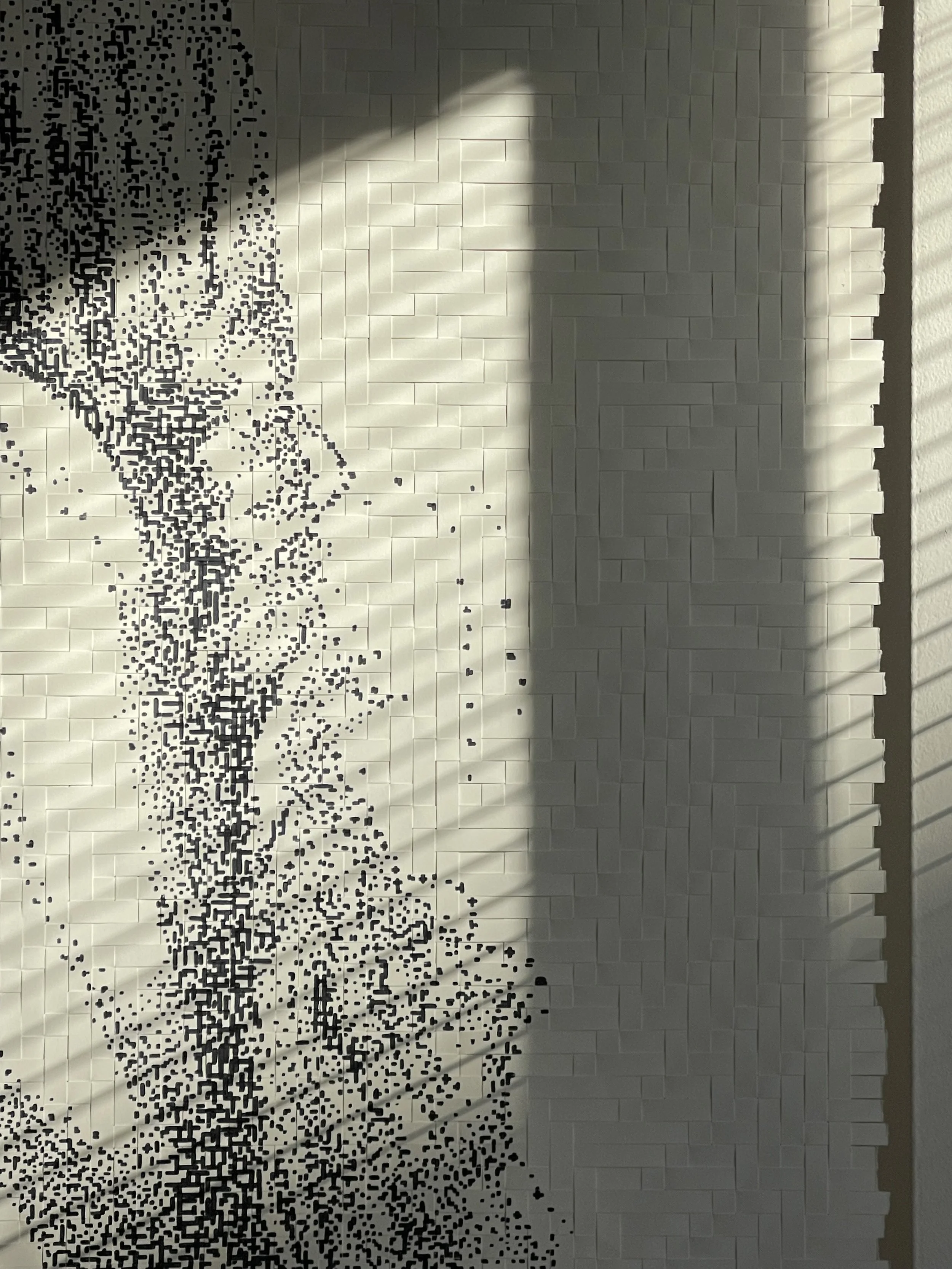 A white textured wall with a vertical black abstract dotted pattern and shadows cast from a window with blinds.