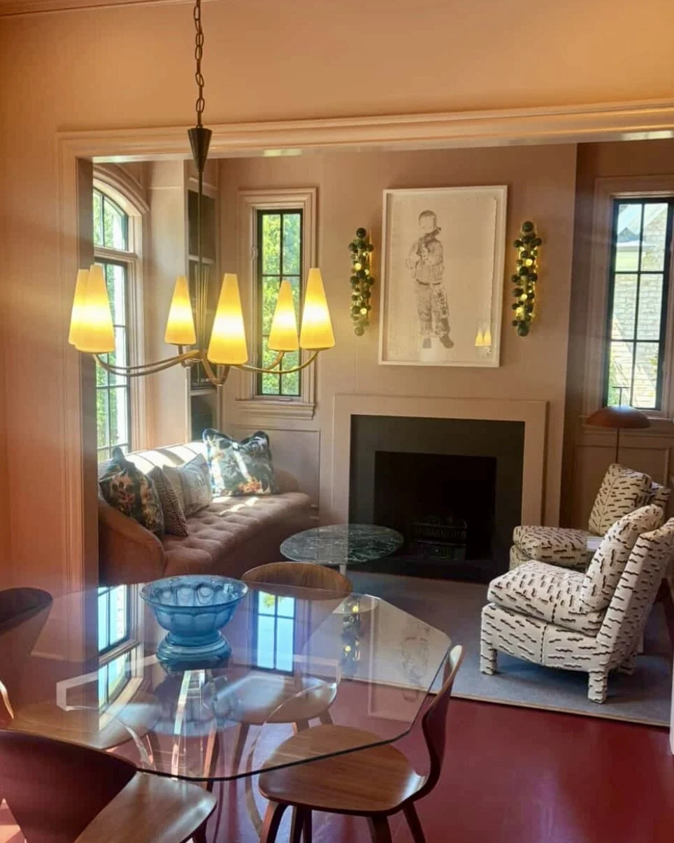 Living room with a glass-top dining table, beige sofa with pillows, patterned armchairs, fireplace, chandelier, and windows with greenery outside.