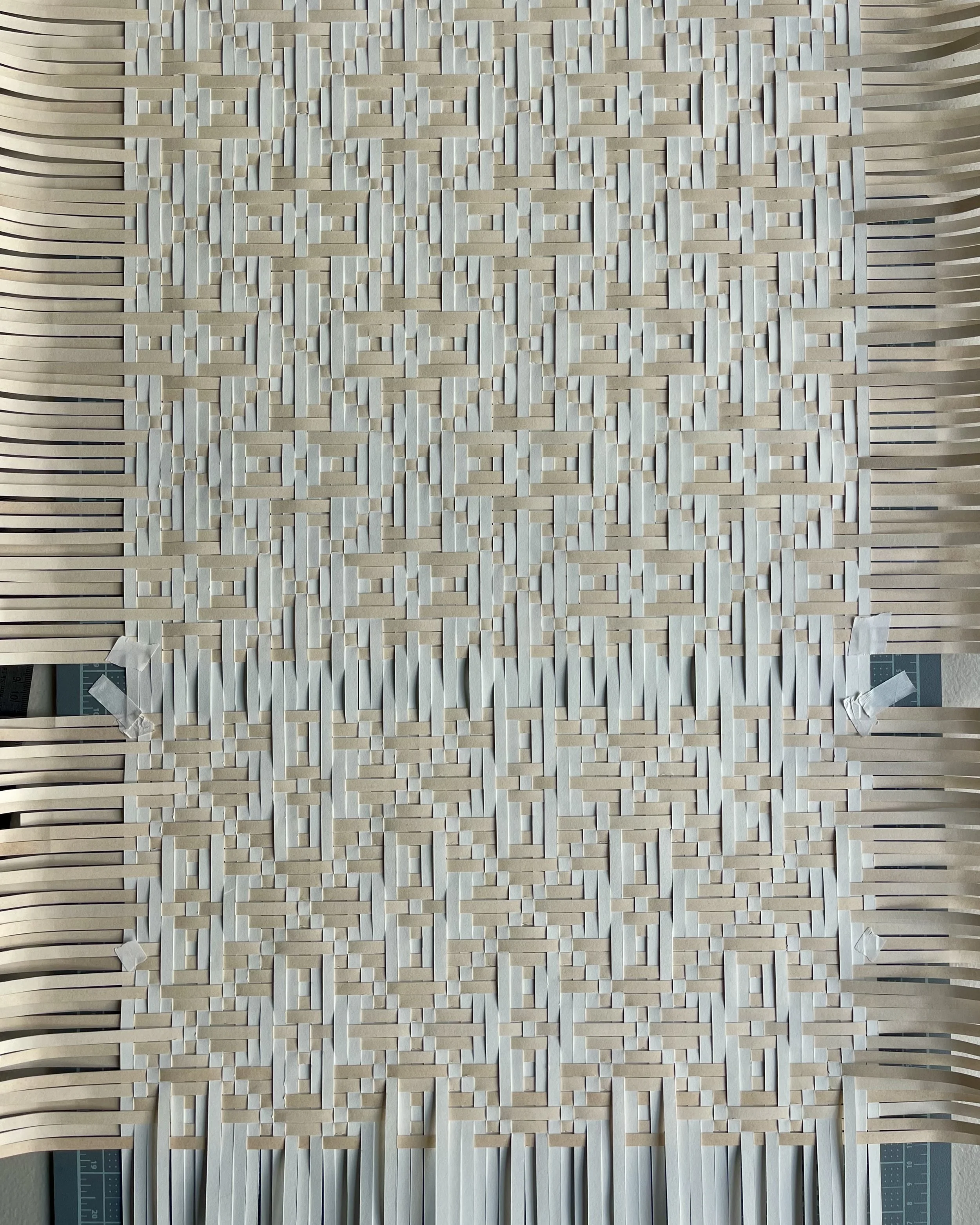 A detailed view of a woven art piece made of cream and white strips of various lengths, creating an intricate geometric pattern with horizontal and vertical elements, secured at the corners with pieces of tape.