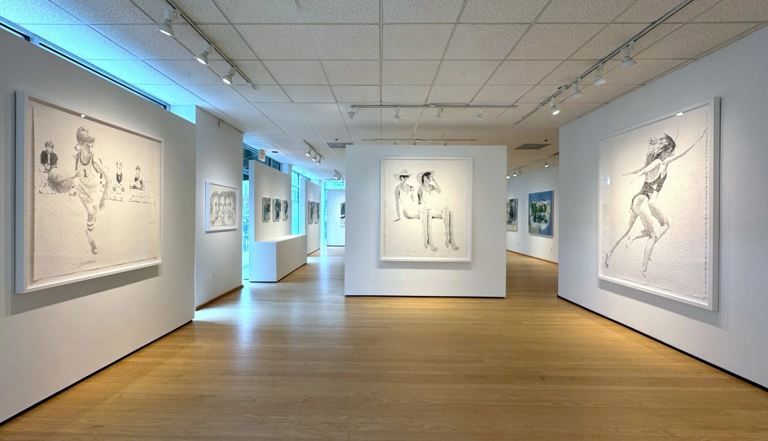 Art gallery with white walls and wooden floors displaying paintings of women, including one of a girl in a swimsuit jumping rope and another sitting on a bench with two other women, illuminated by track lighting.