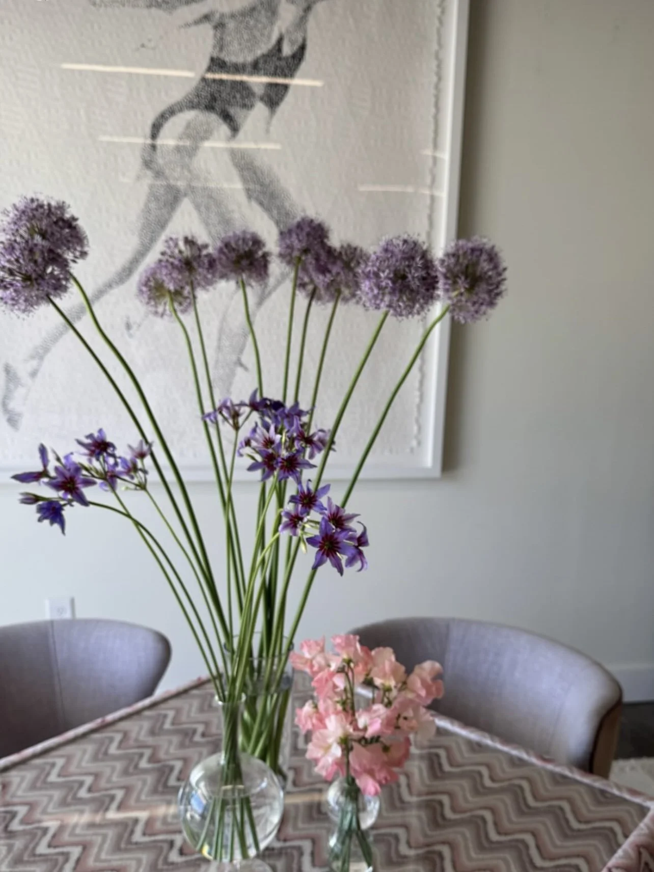 A vase with purple and pink flowers on a table in a room with a large art piece of a woman running on the wall in the background.