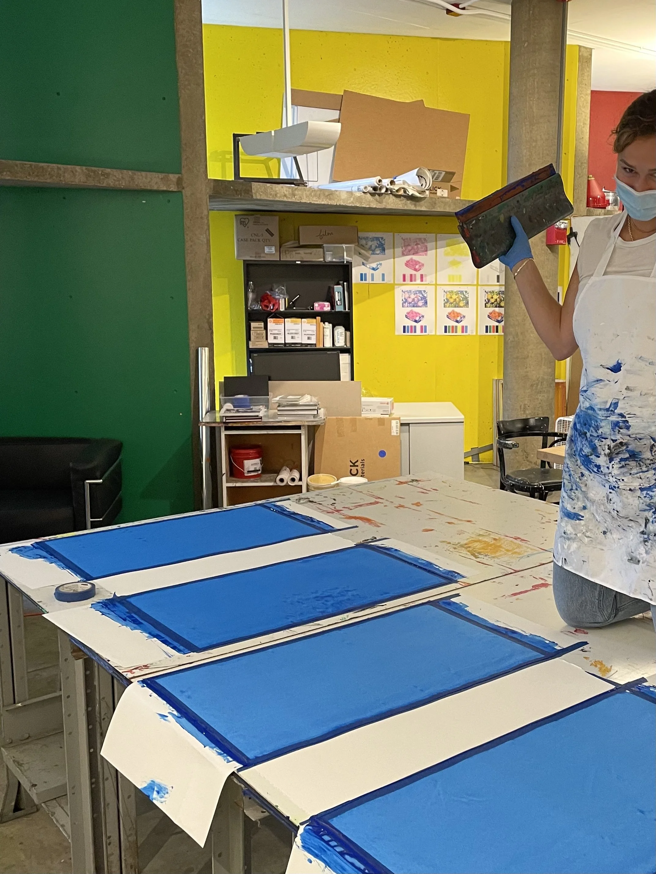 A woman kneeling on a table covered with white paper, printing cyan blue rectangular designs with screen printing. She holds a squeegee and wears a mask, gloves, and a paint-splattered apron.
