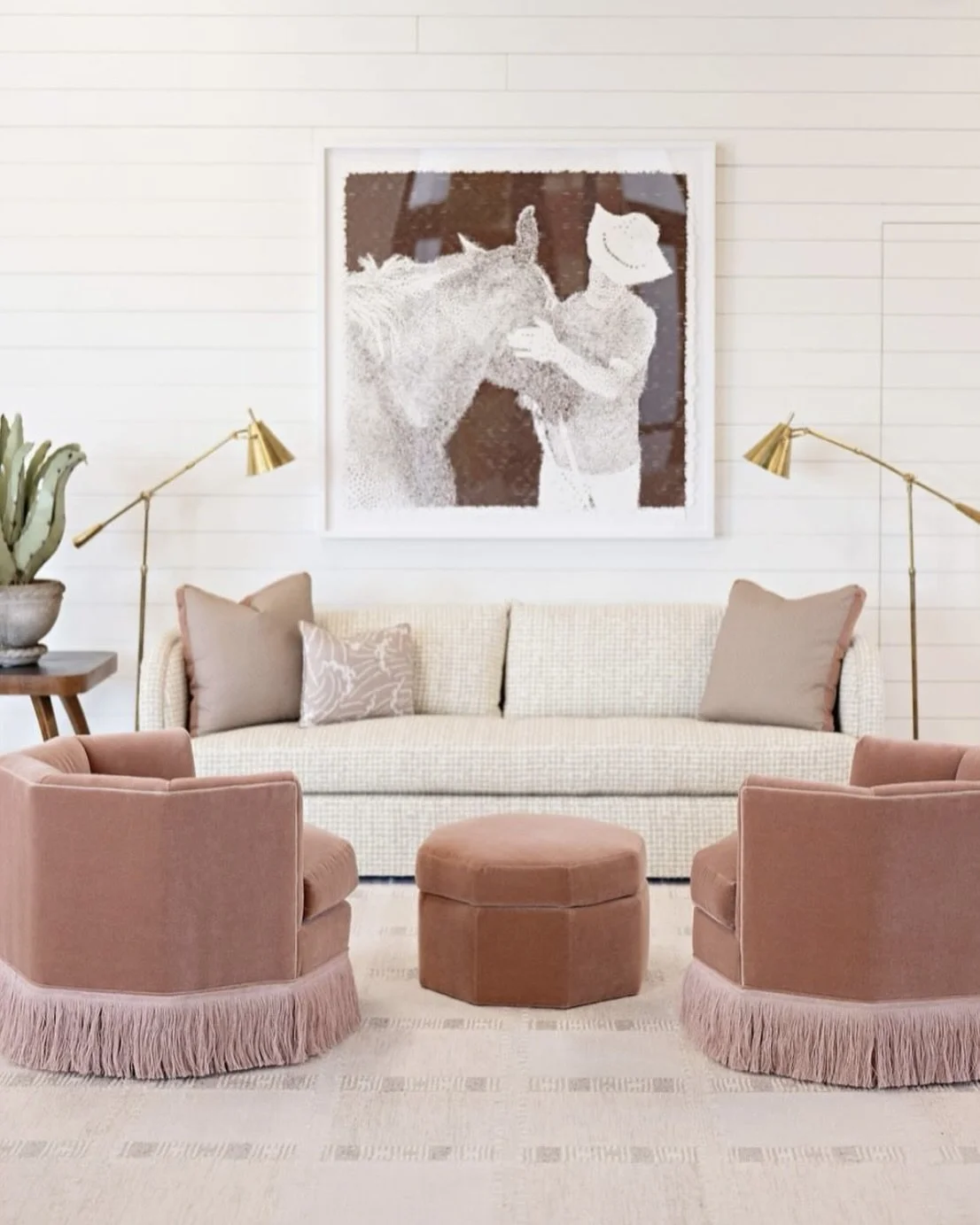 Living room with a white sofa, pink armchairs with fringed skirts, a hexagonal pink ottoman, wall-mounted artwork of a person with a hat and an animal, and gold lamps.