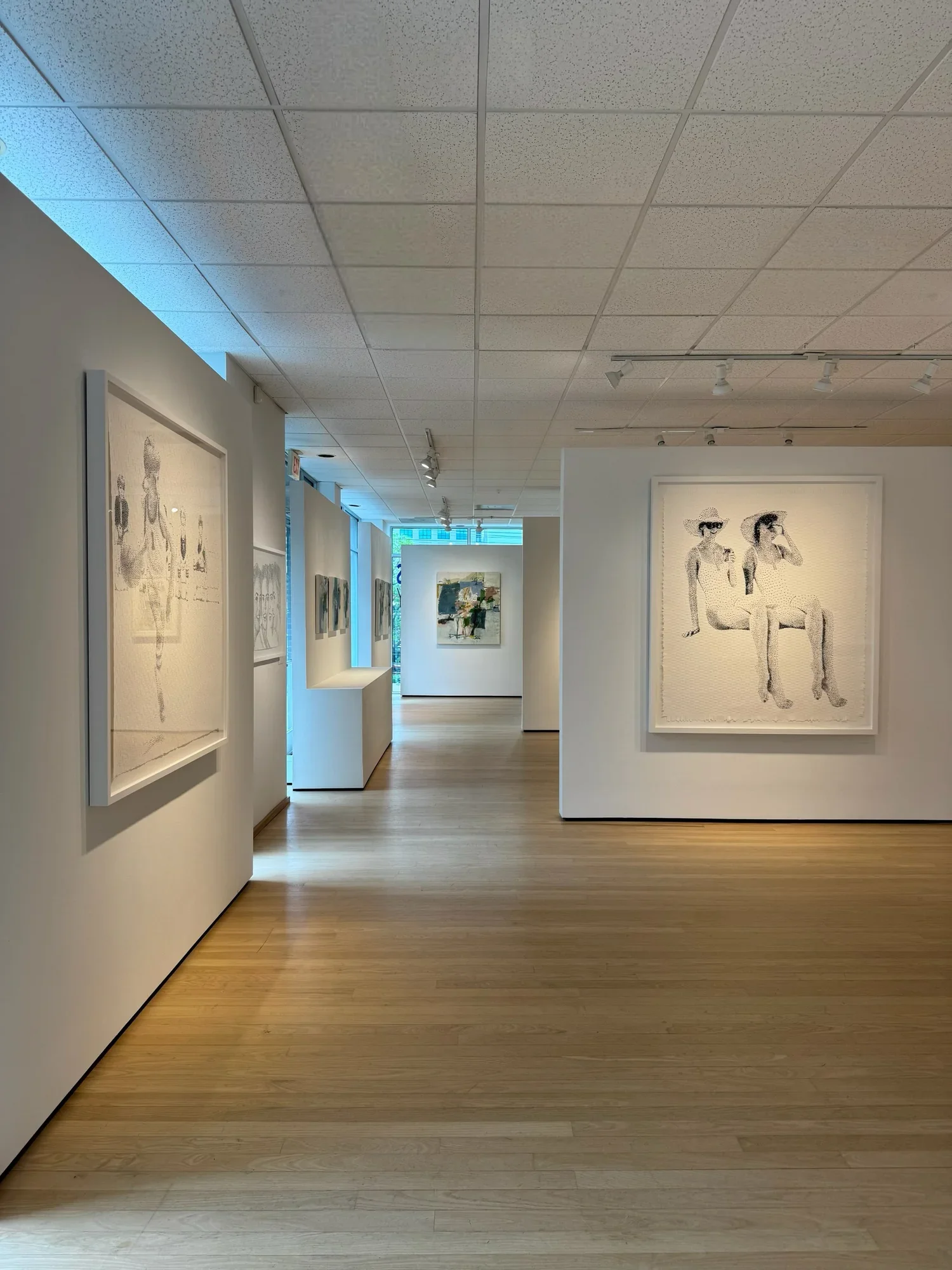 Modern art gallery with white walls displaying various paintings, hardwood floors, and ceiling lights.