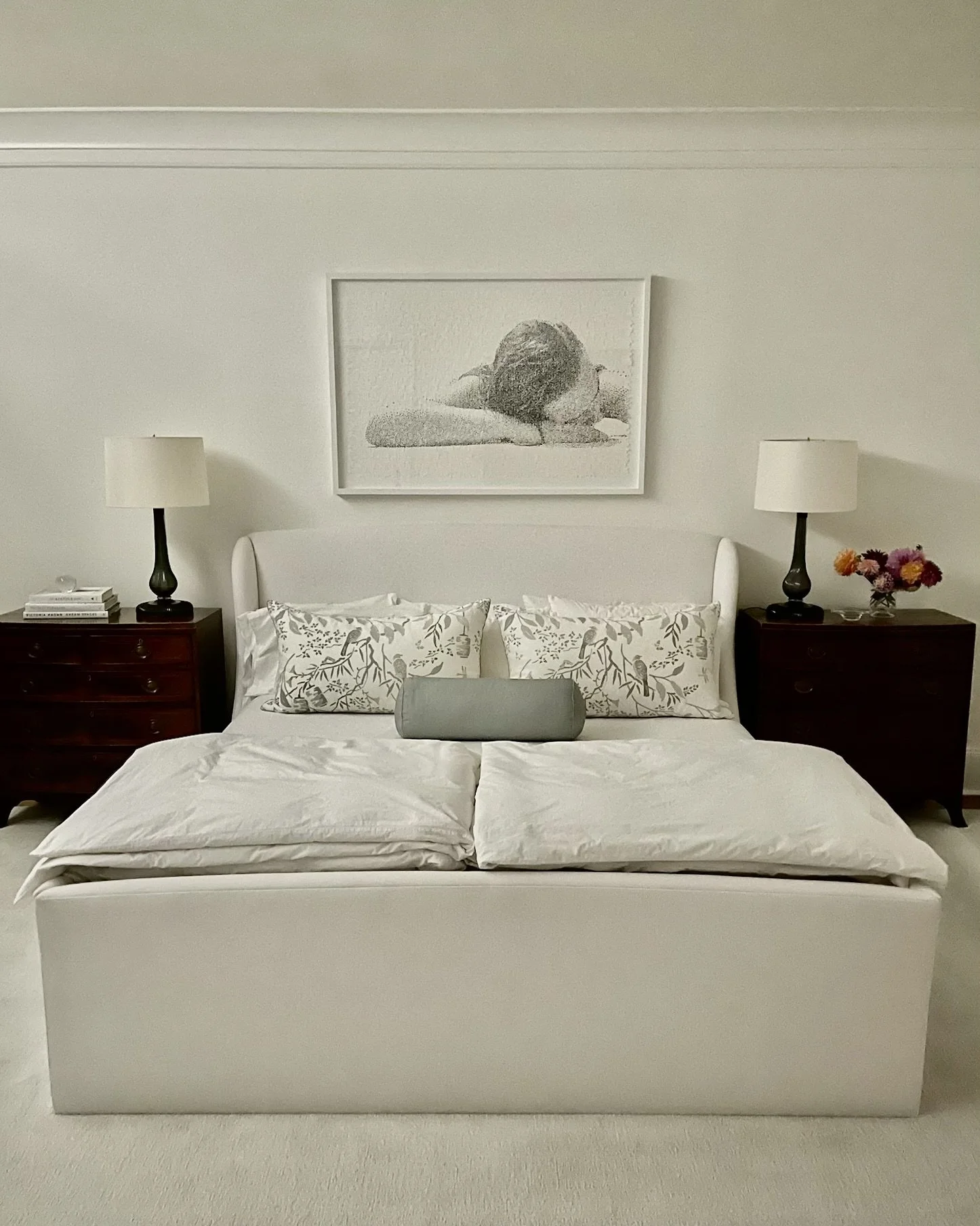 A neatly made bed with white bedding and decorative pillows, centered against a white wall. Above the bed is a black-and-white portrait of a woman resting her head on her arms. Two dark wood nightstands flank the bed, each with a white lamp. The left