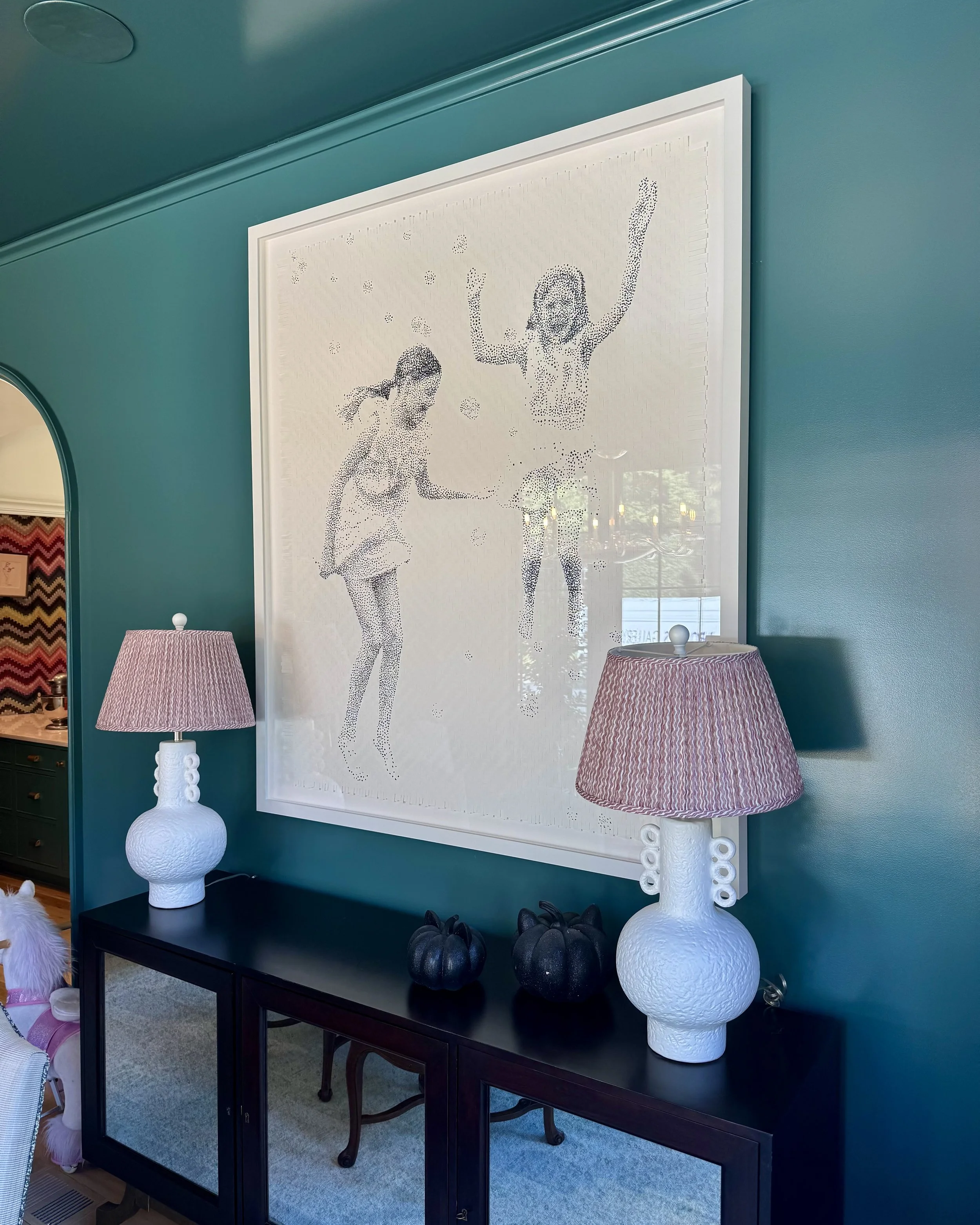 Large white framed artwork of two girls jumping with joy, displayed on a teal wall in a living room. Two white textured table lamps with pink shades are on either side of a black console table, which holds three black pumpkin decorations.
