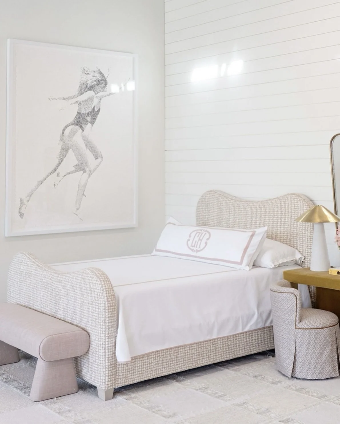 A cozy bedroom with a light-colored bed, decorative pillow, bedside lamp, and artwork of a woman in a swimsuit on the wall.