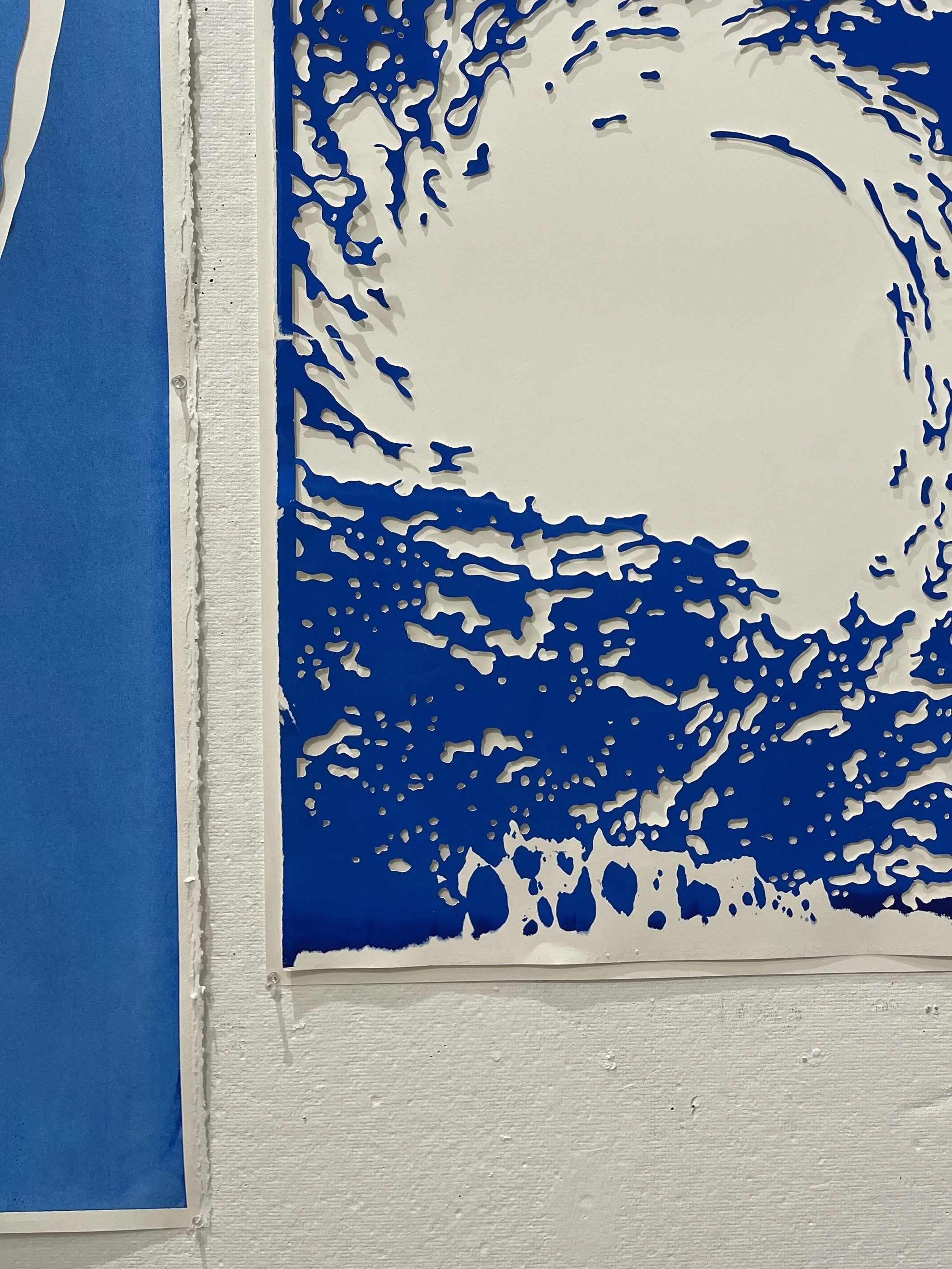 A stencil art of a person surfing a wave, created with blue cutouts on white paper, hanging on a white wall.