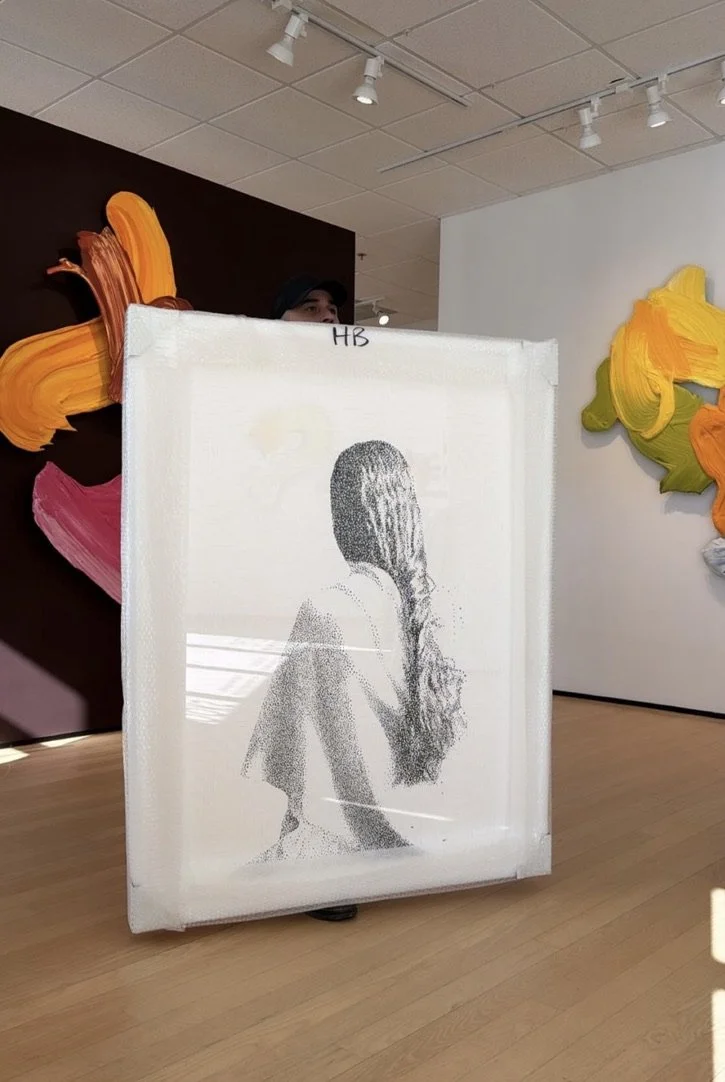 An art installation featuring a black-and-white portrait of a woman with long hair covering her face, created with tiny dots on a semi-transparent canvas. The canvas stands on a wooden floor, with colorful painted abstract shapes in the background.