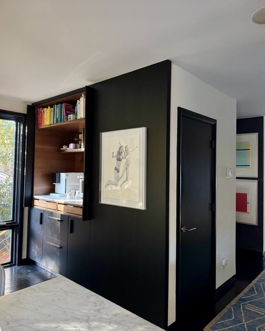 Interior view of a modern apartment with a black and white color scheme, featuring a small kitchen with open shelving, books, and a coffee machine, adjacent to a black door and a wall with framed art.